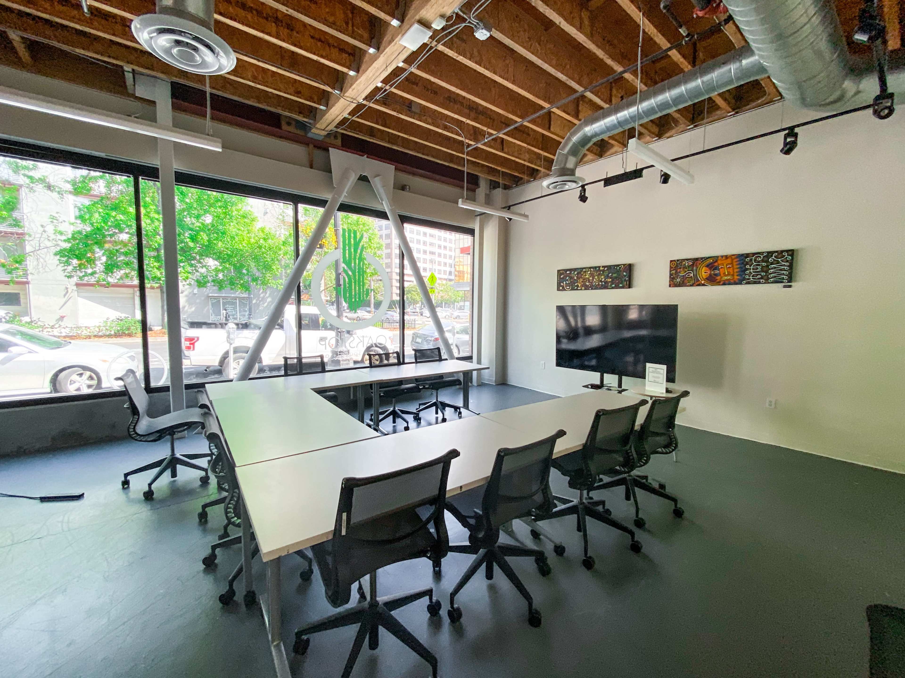 A modern conference room with a large table, several chairs, and a large screen, featuring exposed wooden beams and large windows overlooking a street.