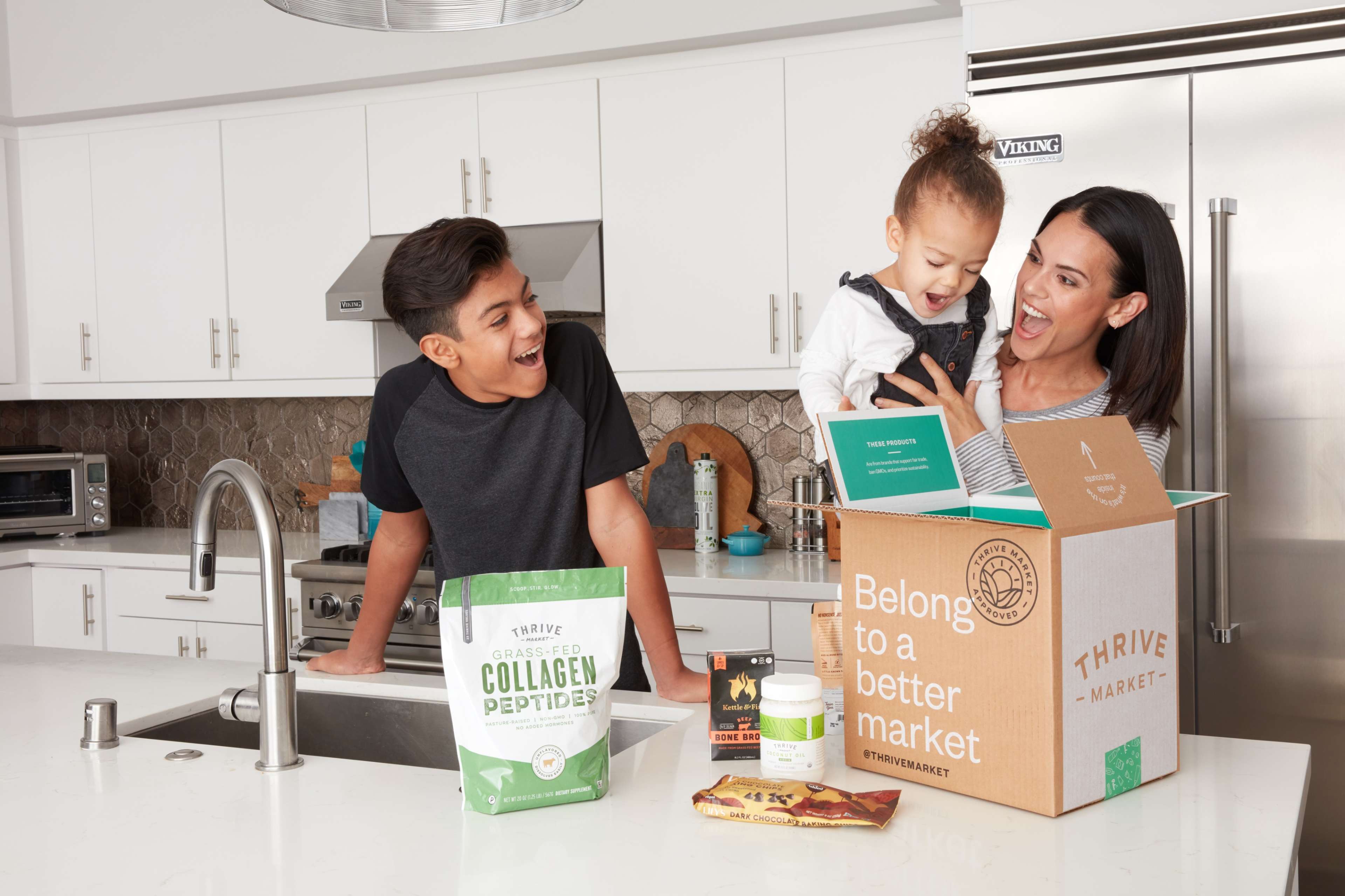 A woman and a boy in a kitchen joyfully unpack a box from Thrive Market while a young child is being held by the woman.