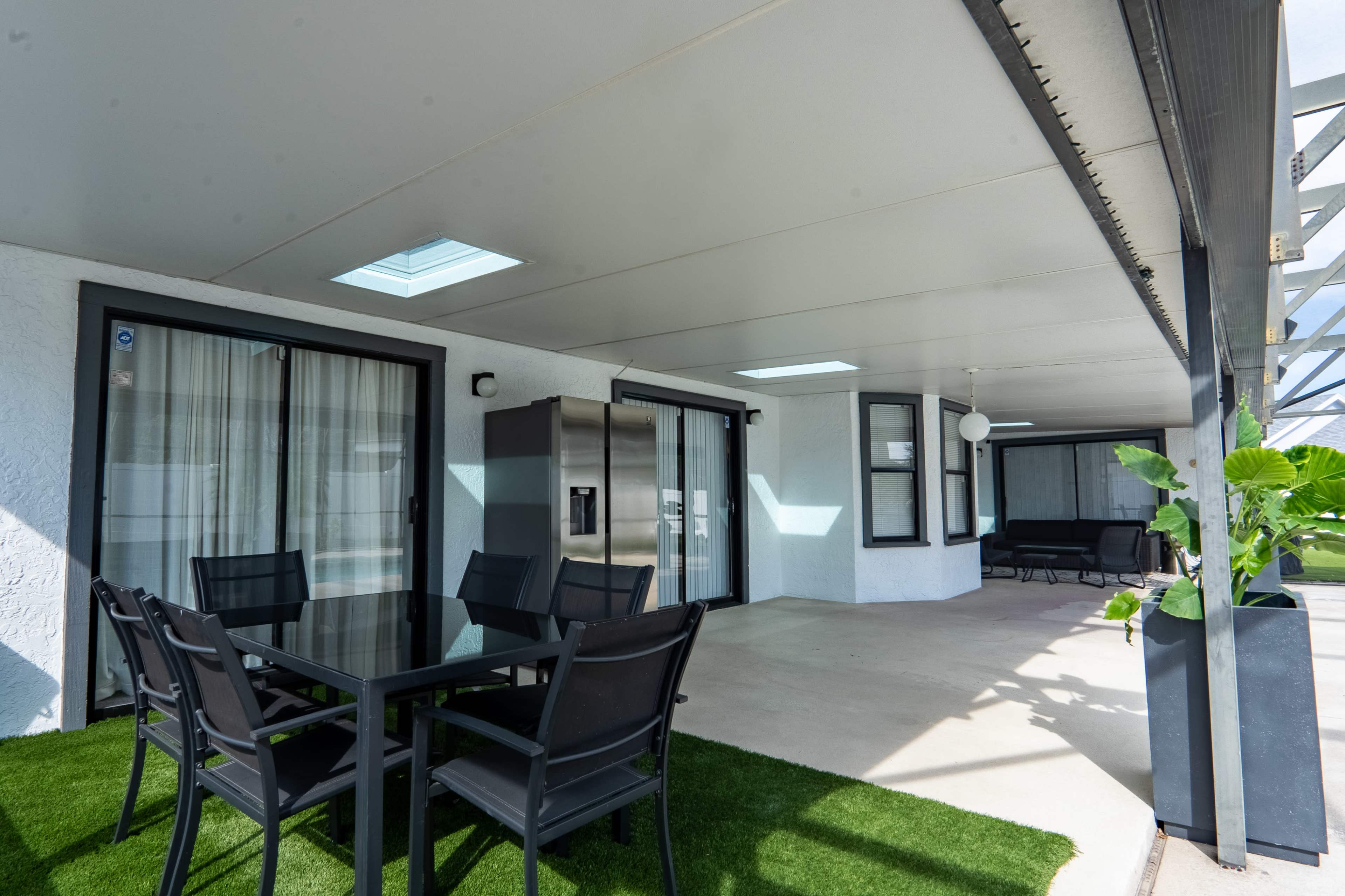 The image shows a covered outdoor patio area with a dining table and chairs, a modern refrigerator, and green artificial grass.