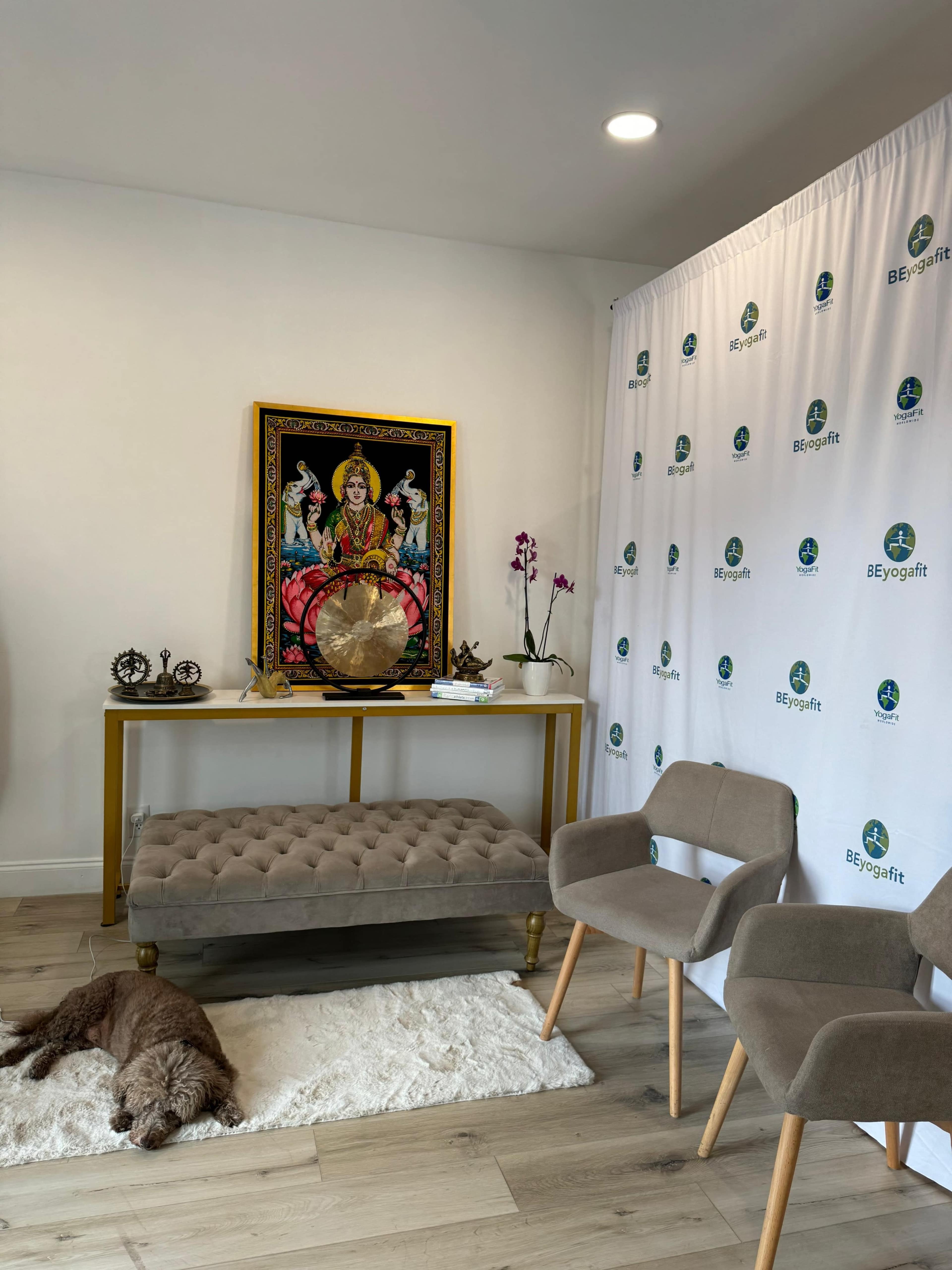 A small room with a decorative table featuring a framed artwork, a globe, and a couch with a rug, alongside two chairs and a small dog resting on the floor.