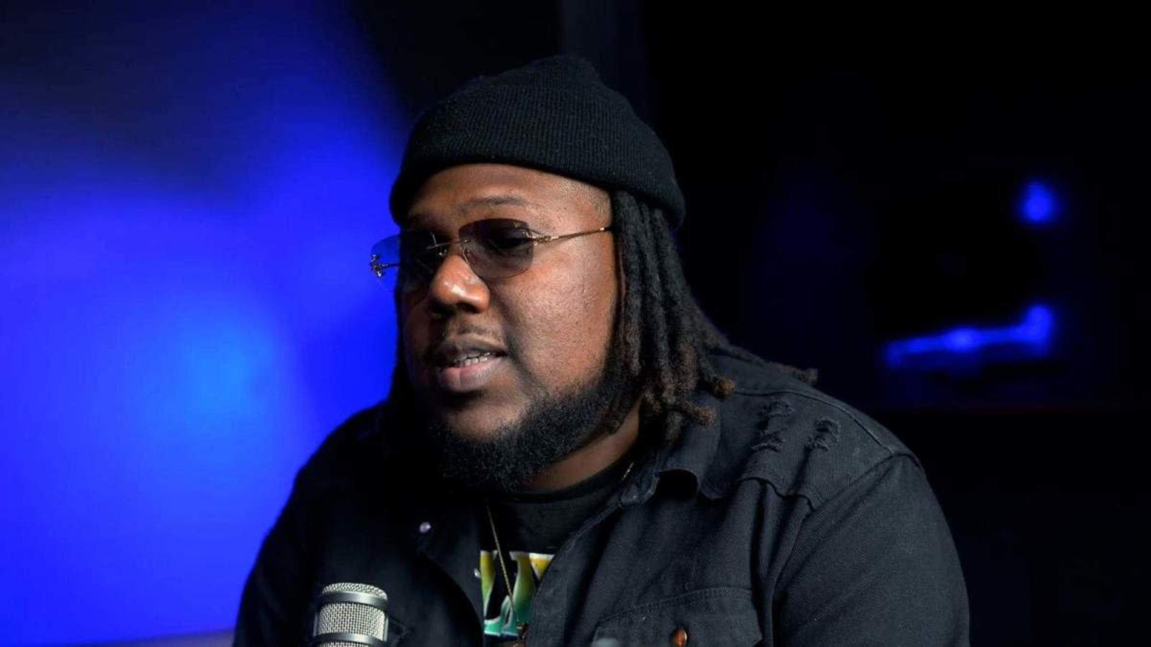 A man with dreadlocks and glasses sits in front of a microphone in a dimly lit setting.