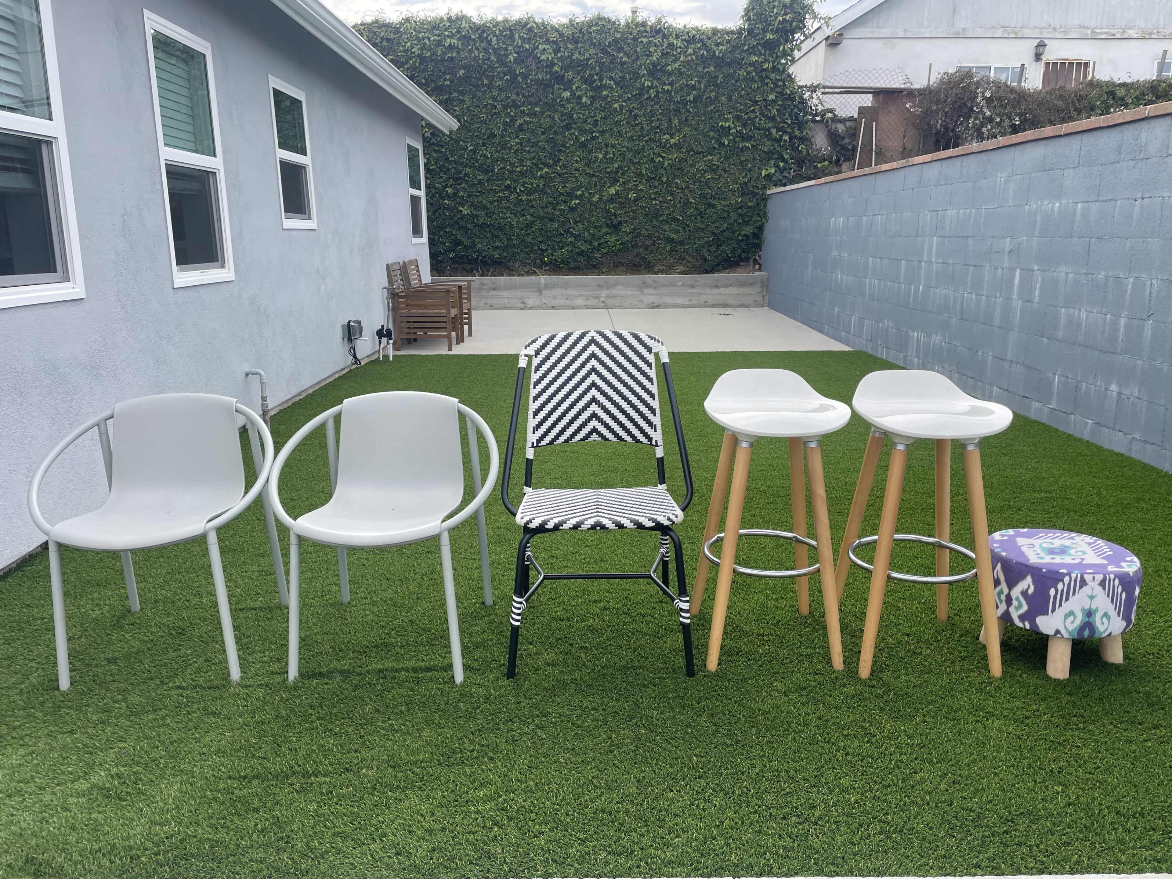 The image shows a row of five outdoor chairs in a green, artificial grass area behind a house, with a gray wall and a concrete patio in the background.