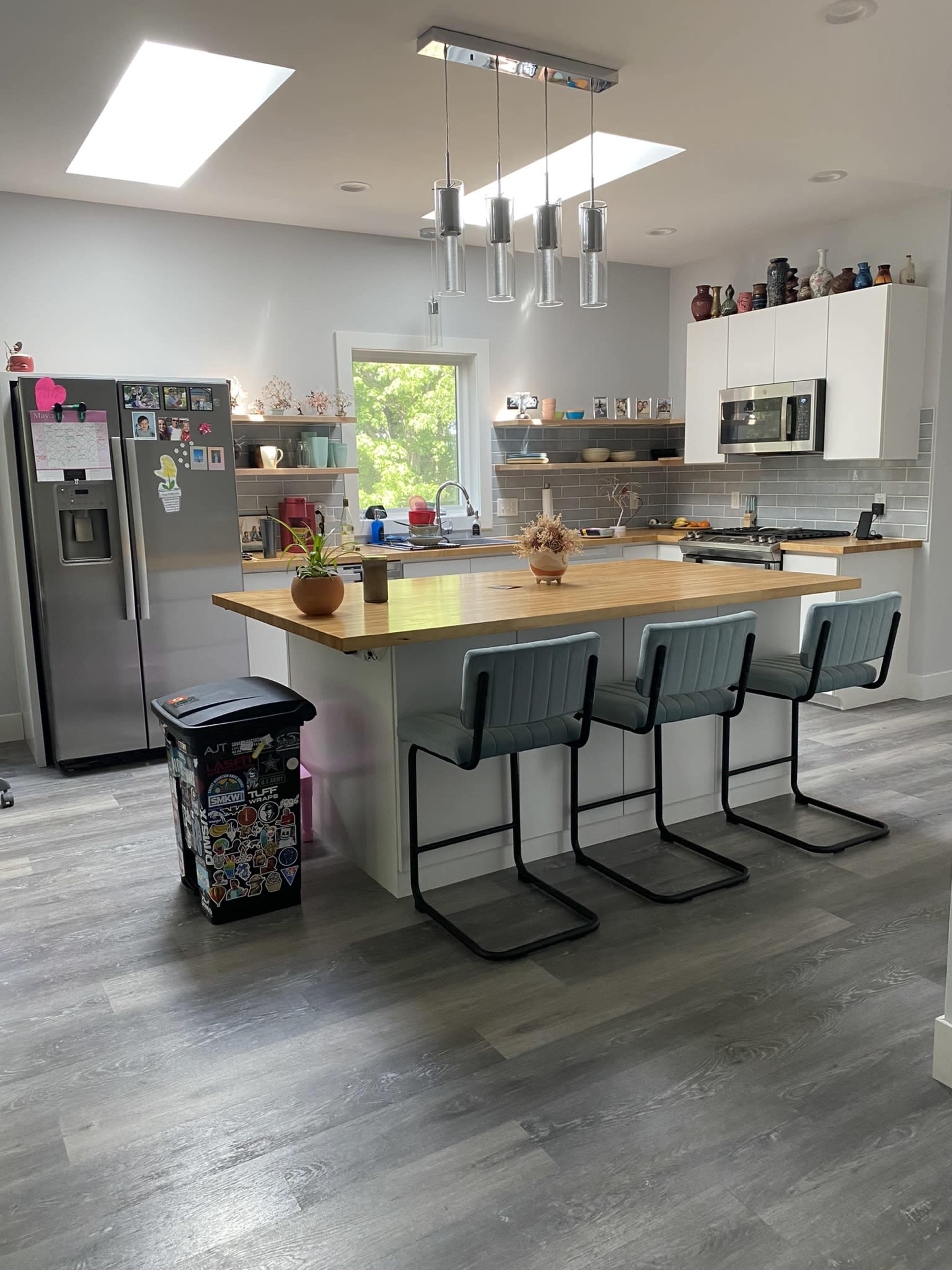 A modern kitchen features a central island with three bar stools, stainless steel appliances, and various decorative items on shelves.