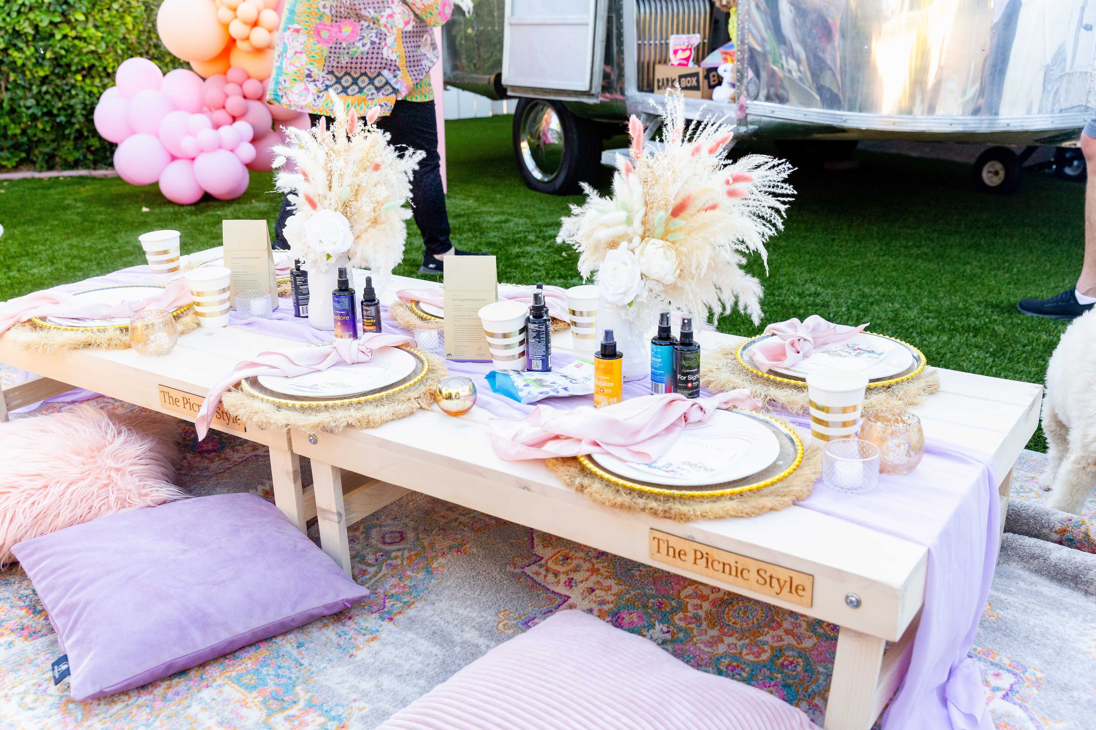 A decorated picnic table with elegant table settings and decorative elements is set up on green grass, surrounded by plush pillows and a shiny trailer in the background.