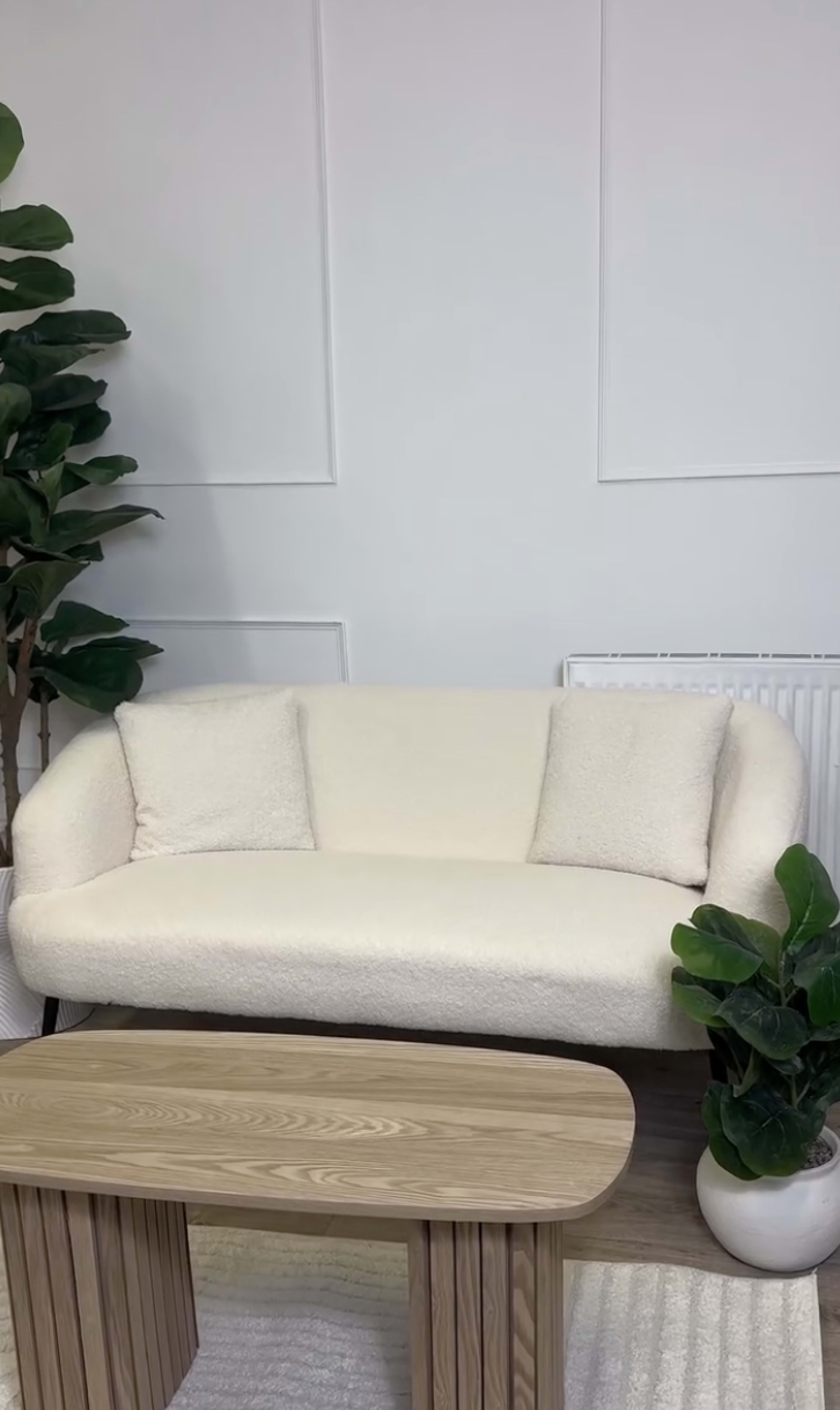 The image shows a light-colored sofa with two cushions, positioned next to a wooden coffee table and surrounded by potted plants in a minimalistic room.