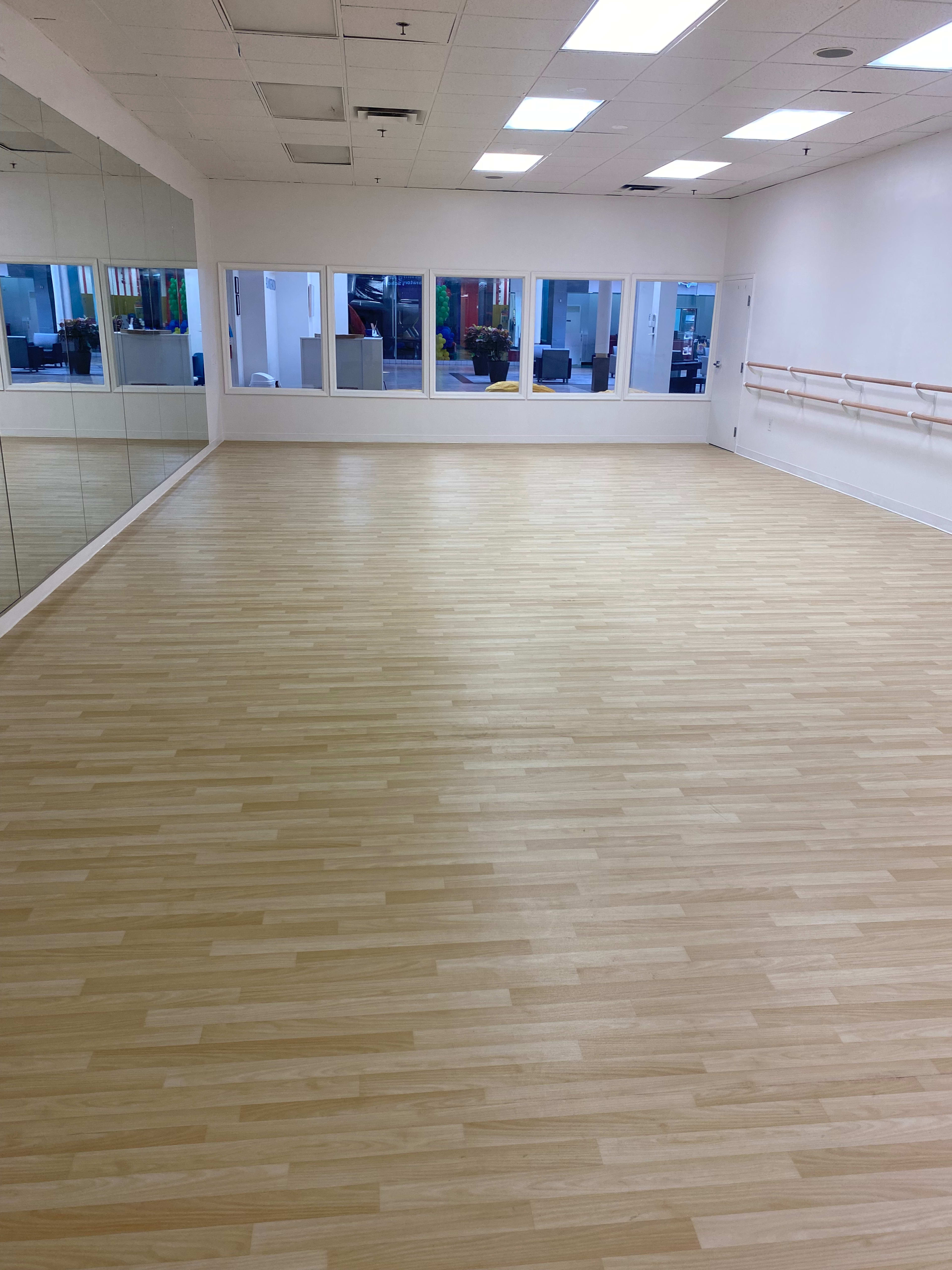 A spacious dance studio with wooden flooring, large mirrors on one wall, and bright natural light coming through the windows.