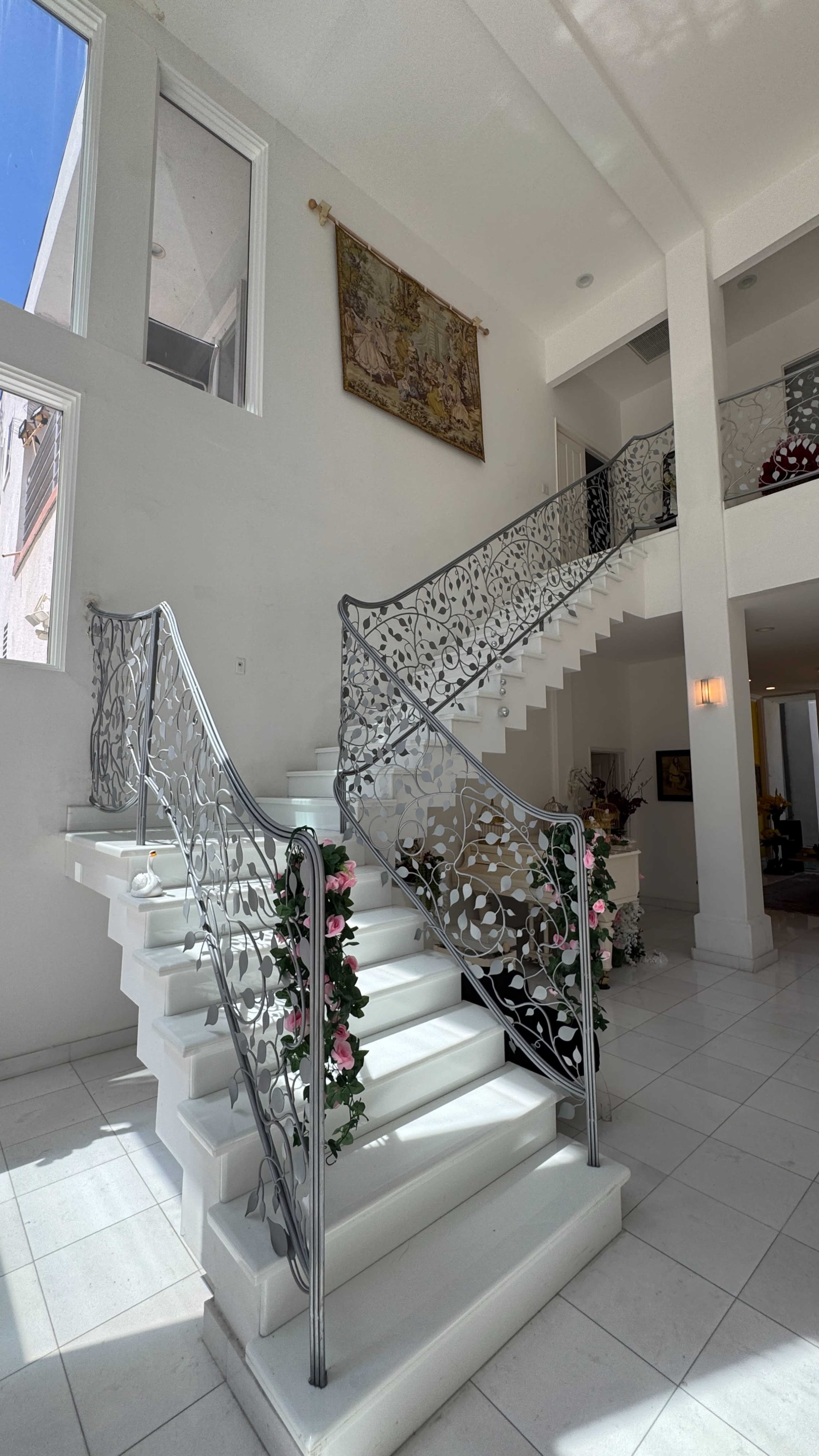 The image shows a bright, spacious foyer featuring two curved staircases with decorative metal railings and floral arrangements along the steps.