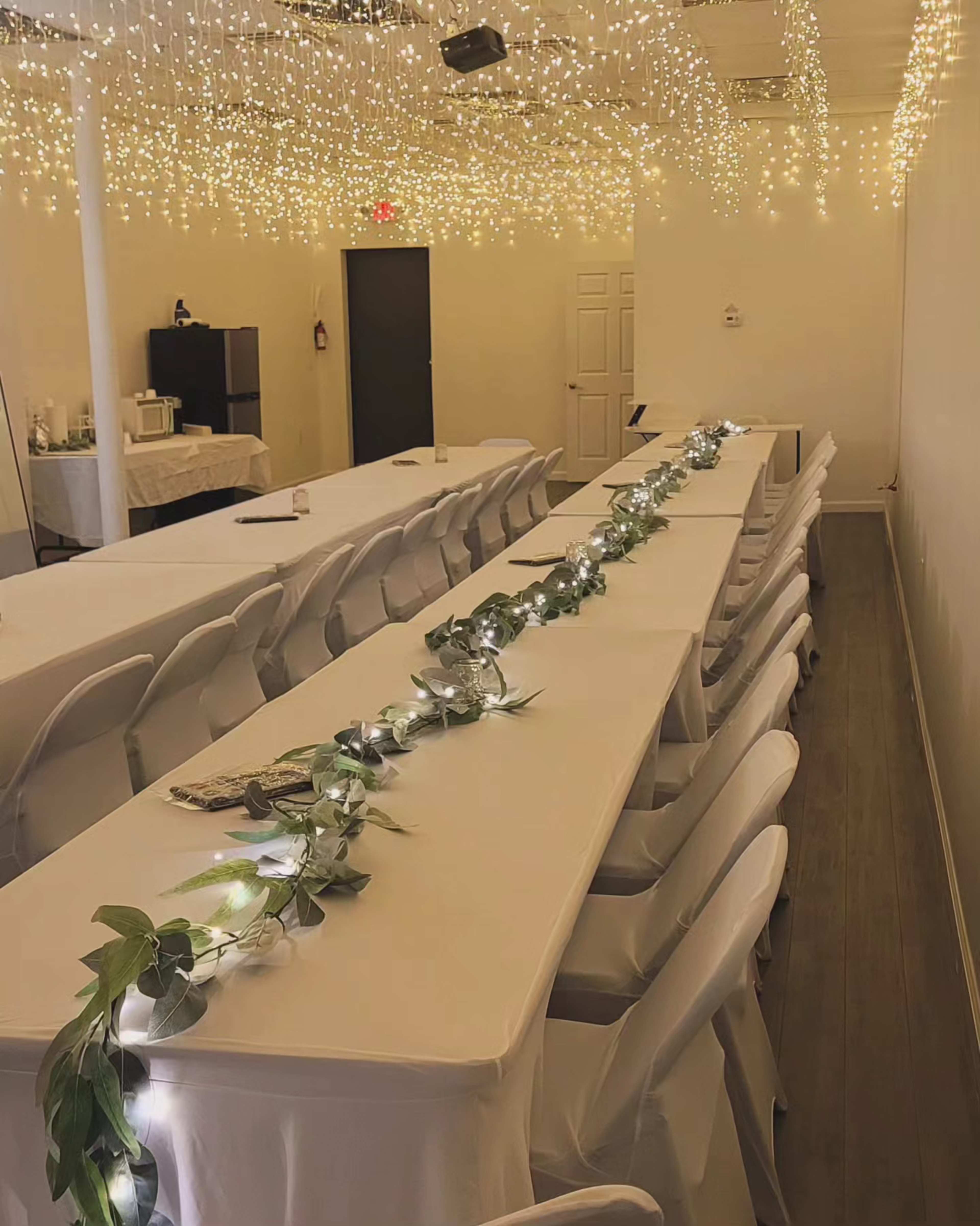 A long banquet table is arranged in a well-lit room, adorned with a greenery and light garland, under a ceiling of strings of lights.