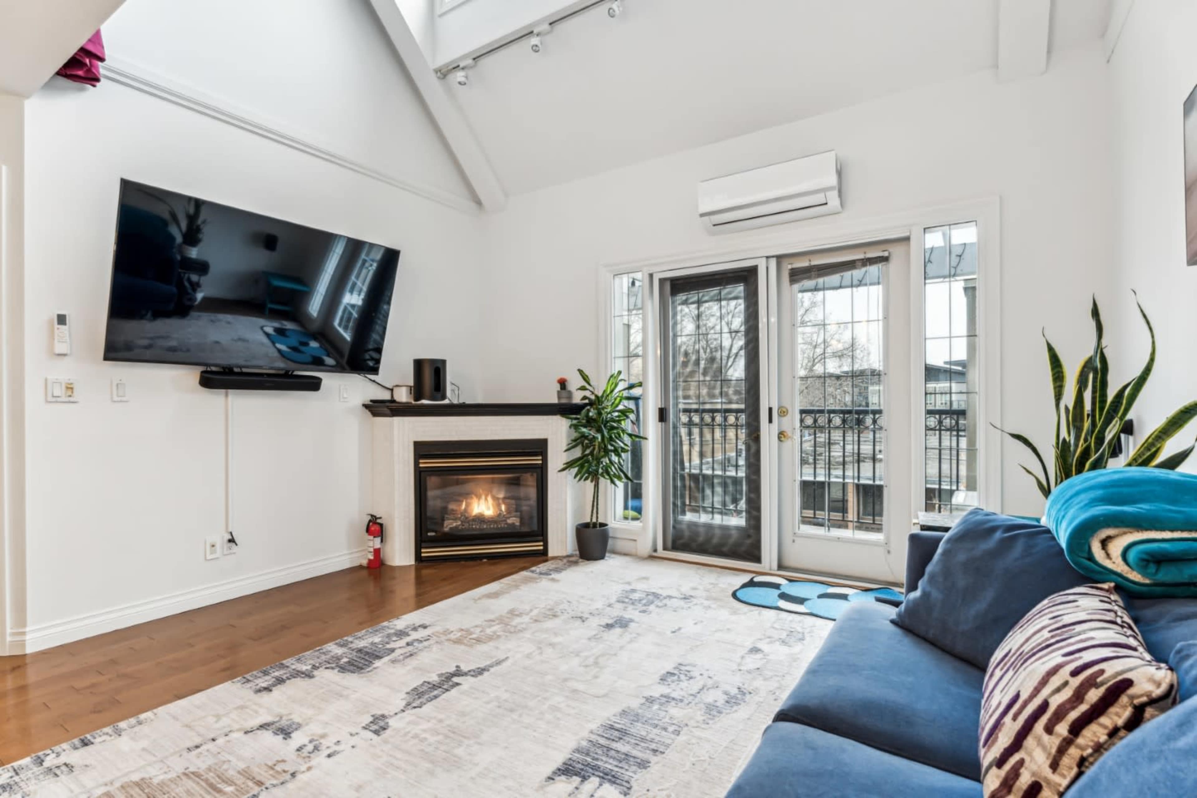 The image shows a modern living room featuring a blue sofa, a large TV mounted on the wall, a fireplace, and sliding glass doors leading to a balcony.