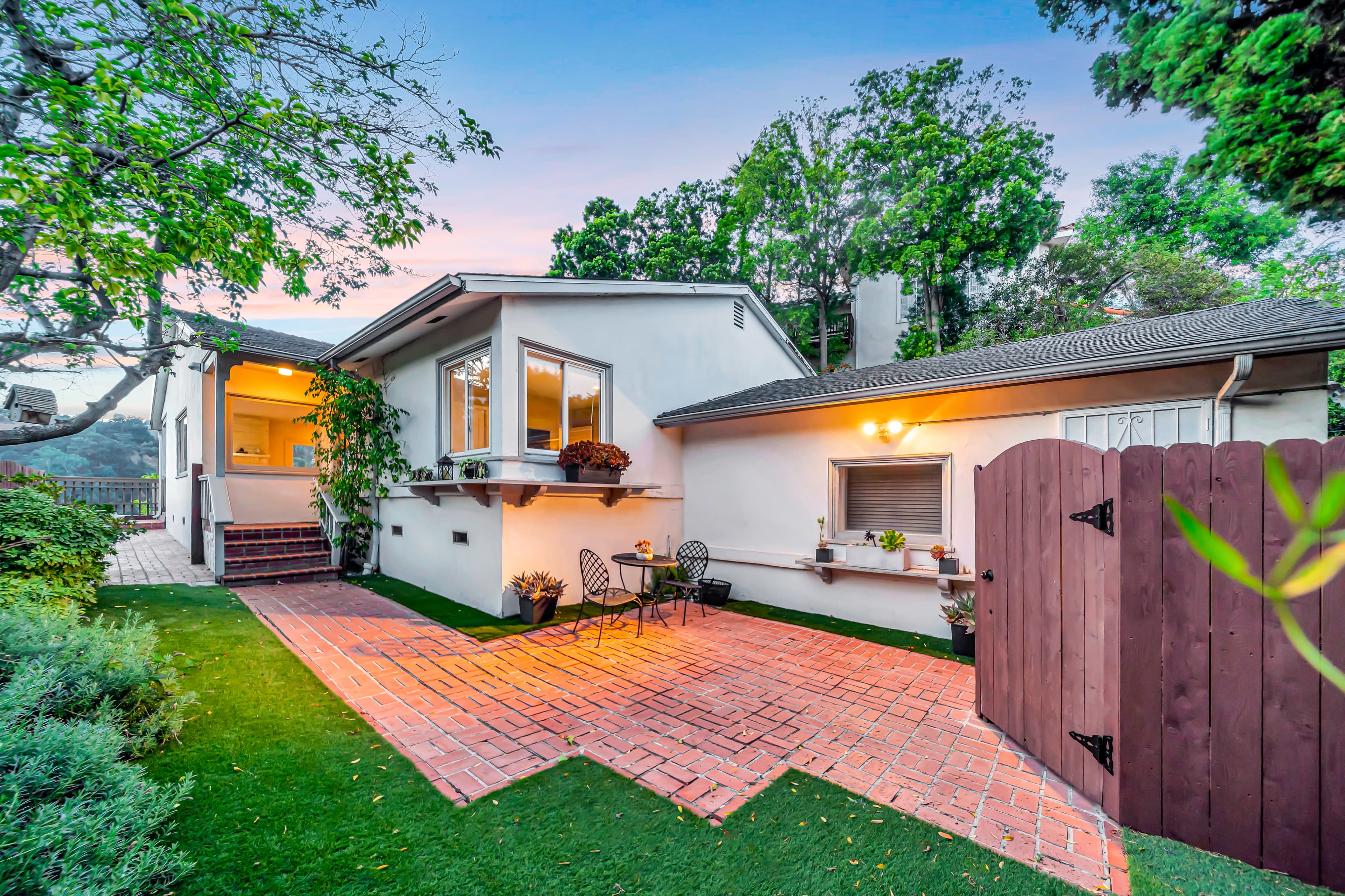 A modern house with a brick pathway and a small garden is surrounded by trees and features a wooden gate.