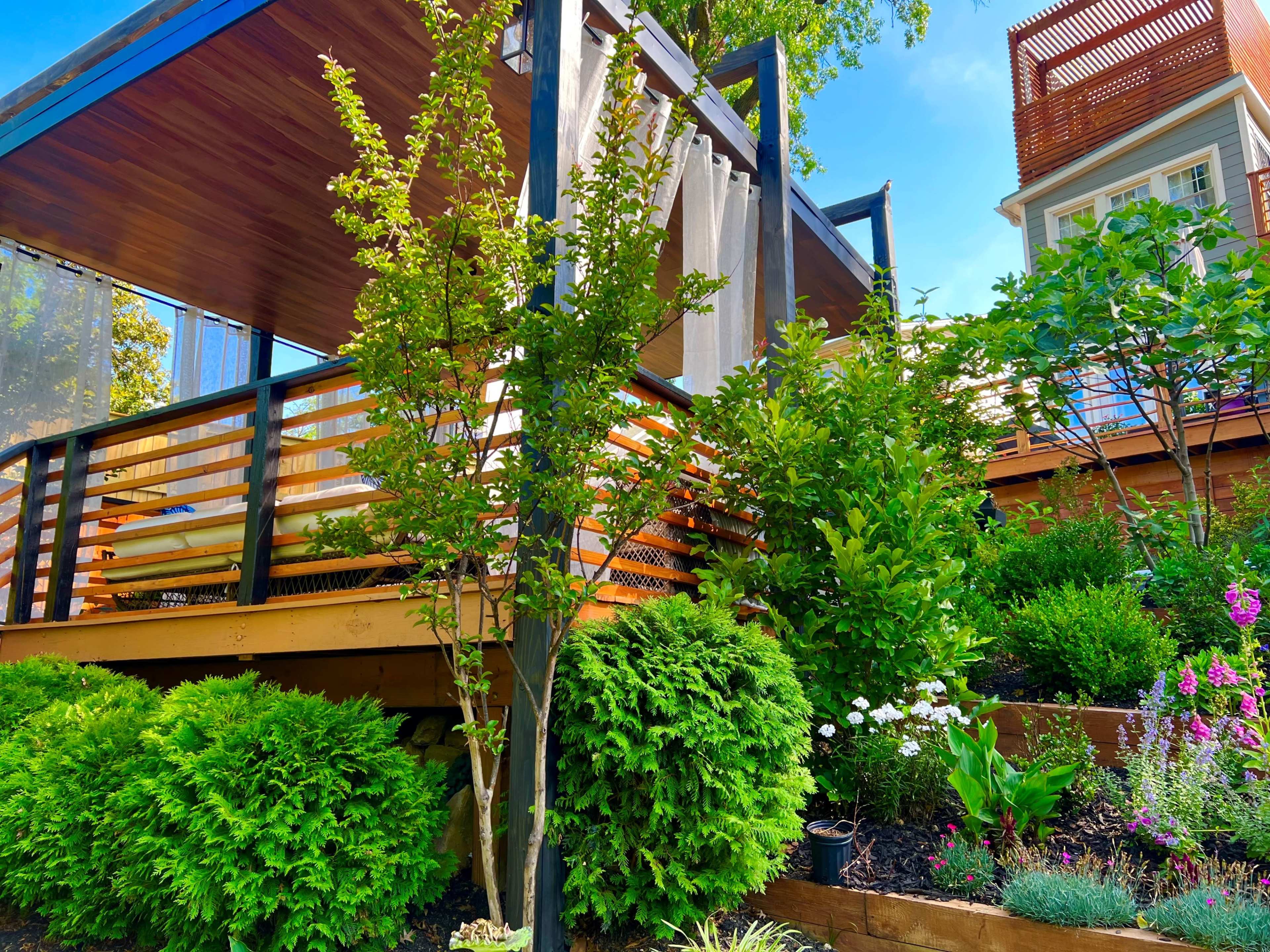 A wooden deck elevated on stilts, surrounded by lush greenery, including bushes and flowers, with a house partially visible in the background.