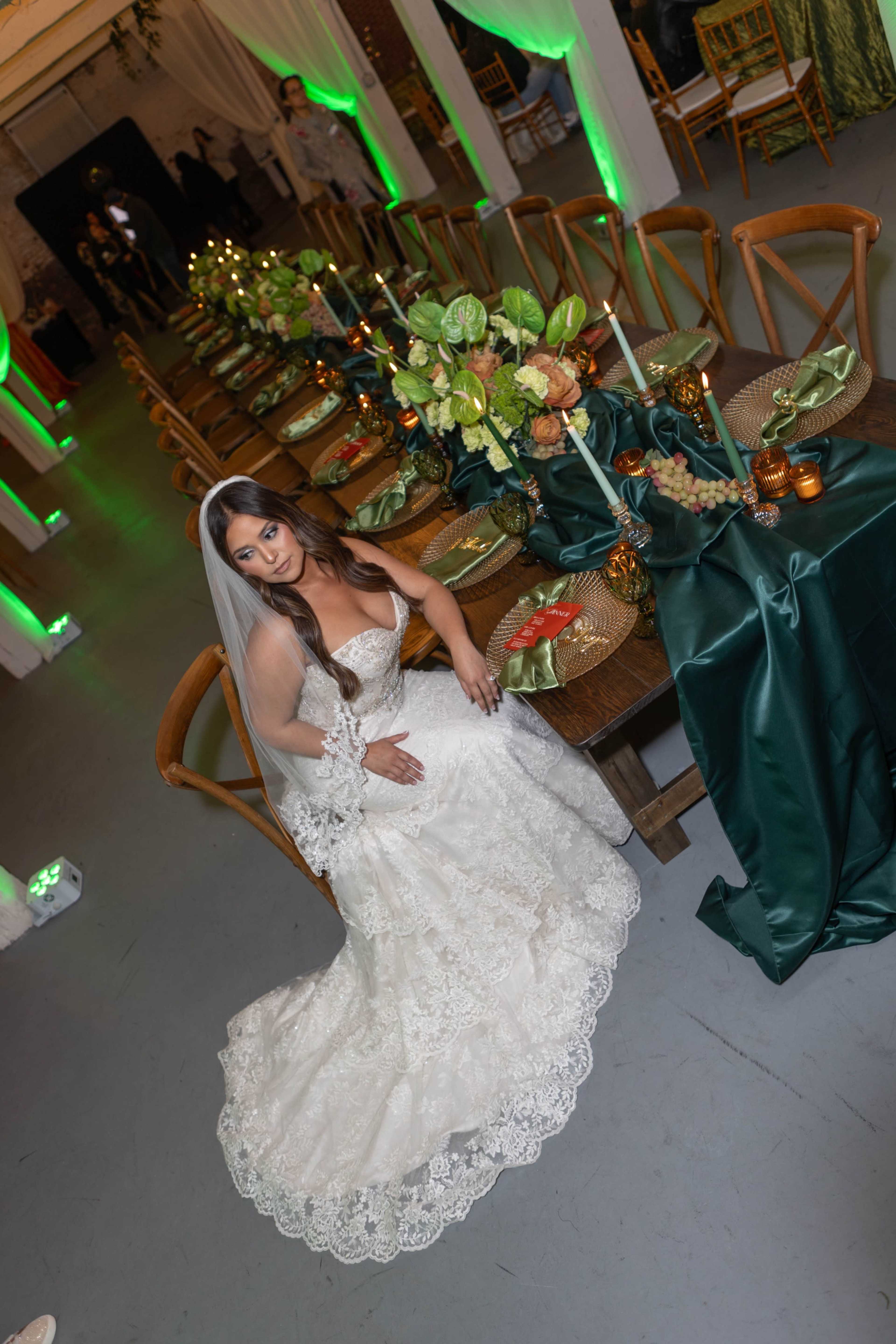 A bride in a white gown sits elegantly on the floor near a long, decorated dining table set with green and gold accents.