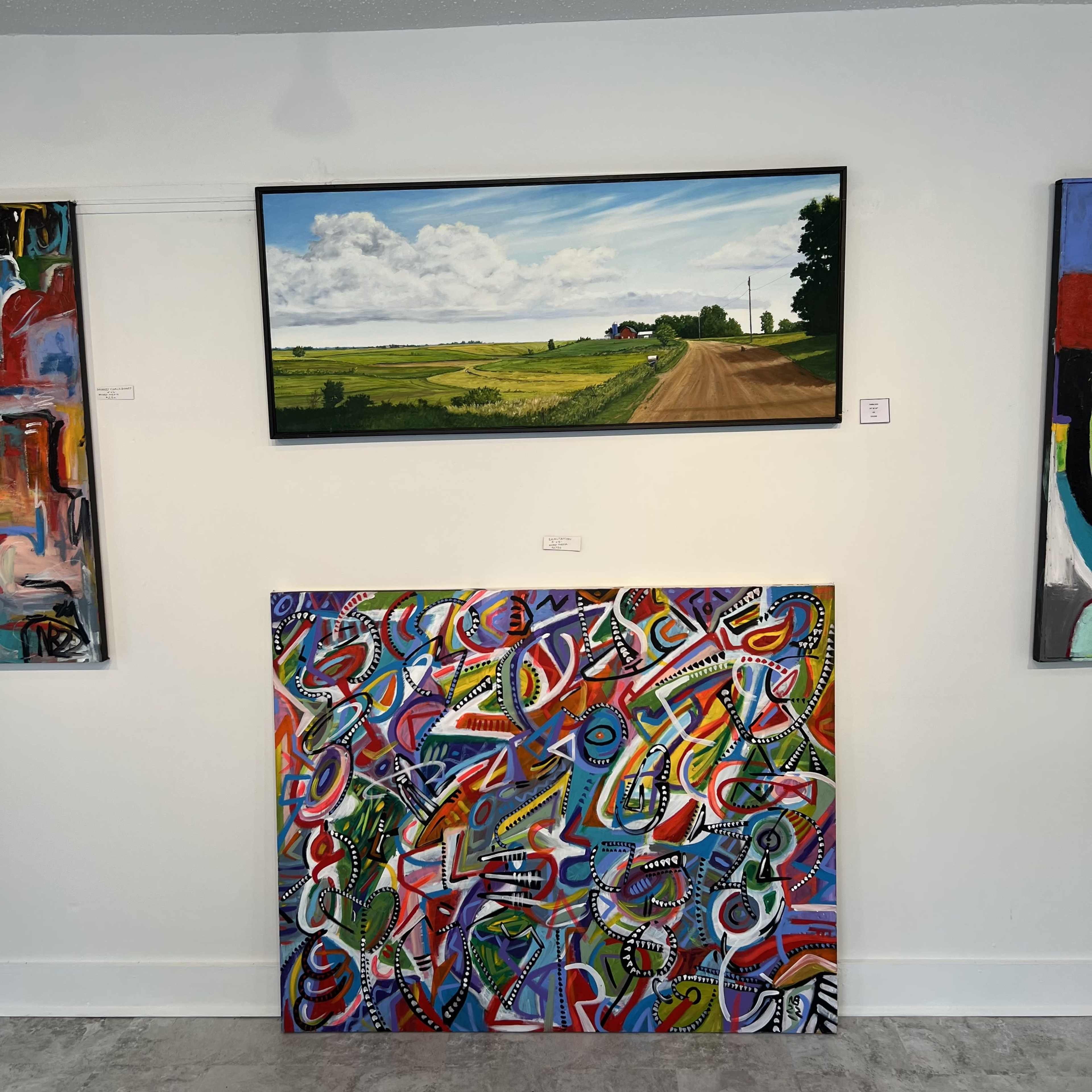The image features a gallery wall displaying two artworks: a horizontal landscape painting above depicting a serene rural scene and a colorful abstract painting below.