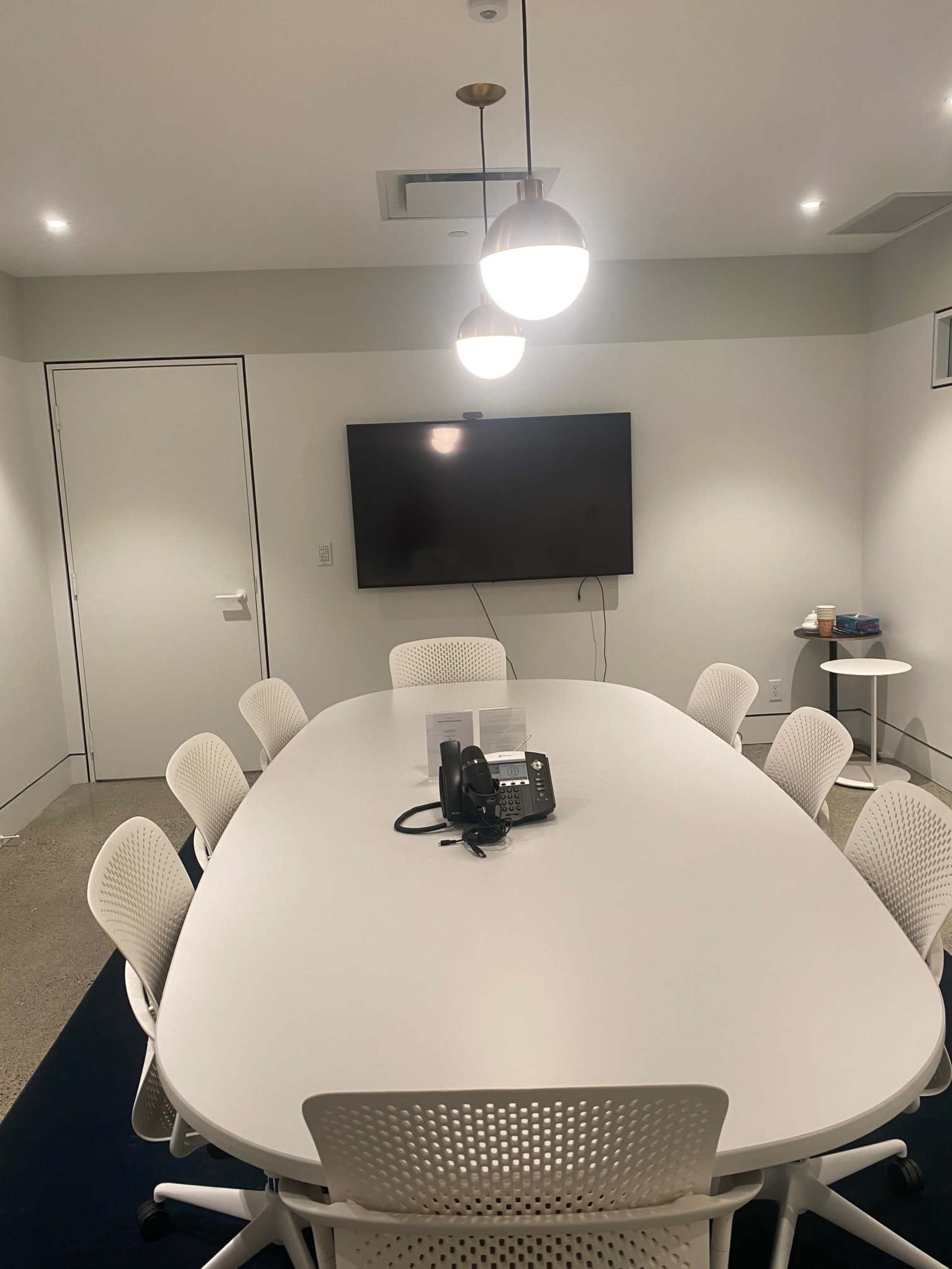 A conference room features a large oval table surrounded by white chairs, a wall-mounted television, and a telephone at the center.