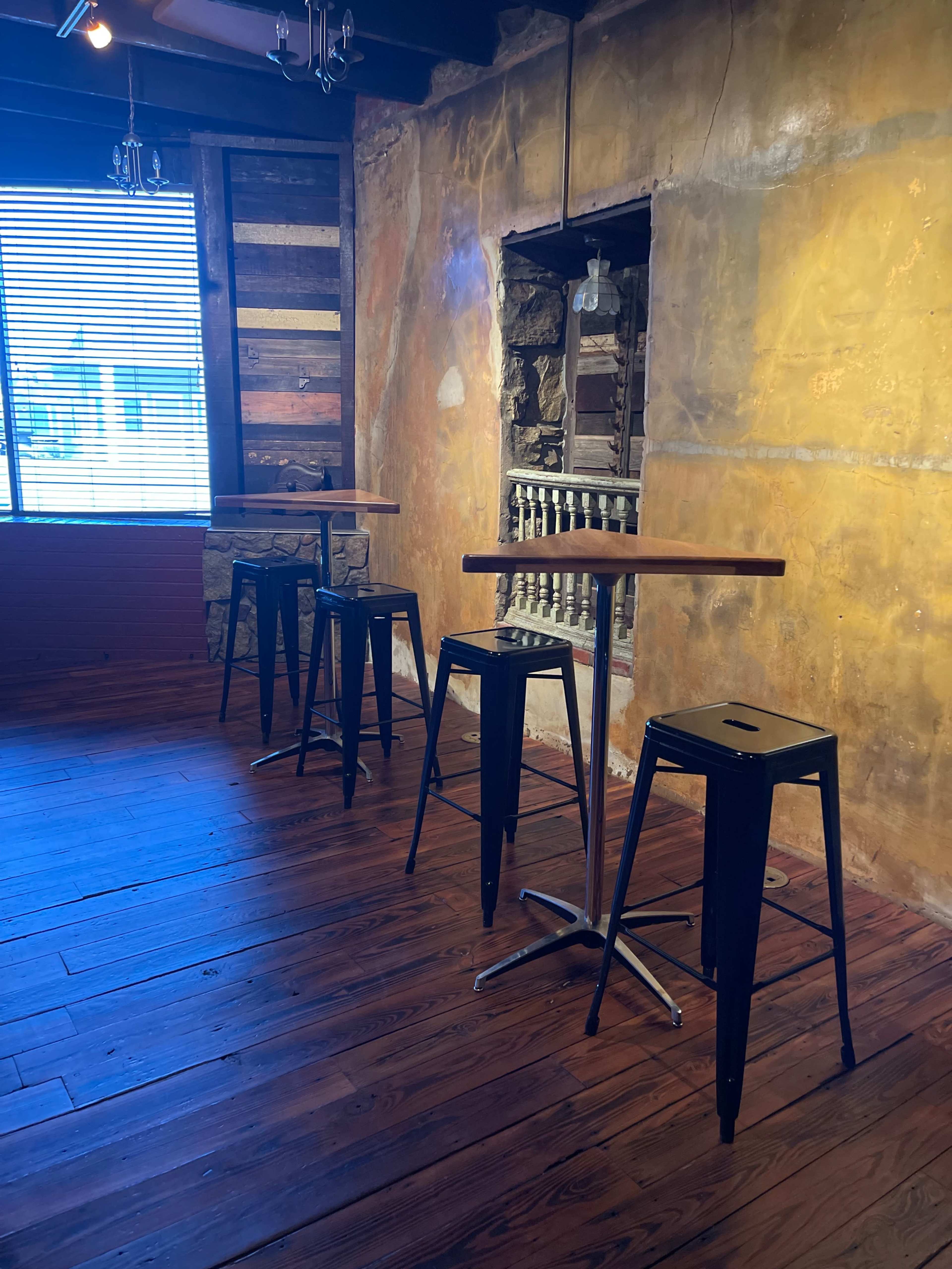 A corner of a room features several black bar stools arranged around tall wooden tables, with a textured wall painted in warm tones and natural light coming through a window.