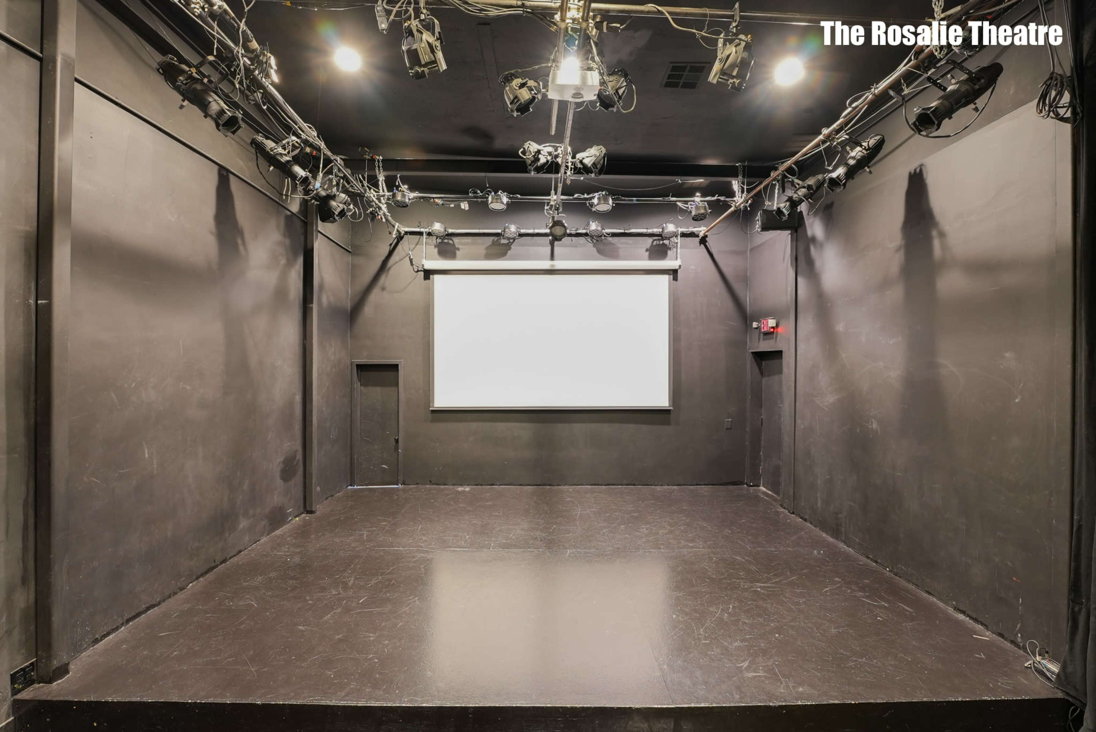 The image shows a dimly lit black box theater with a blank screen at the front and overhead stage lighting.