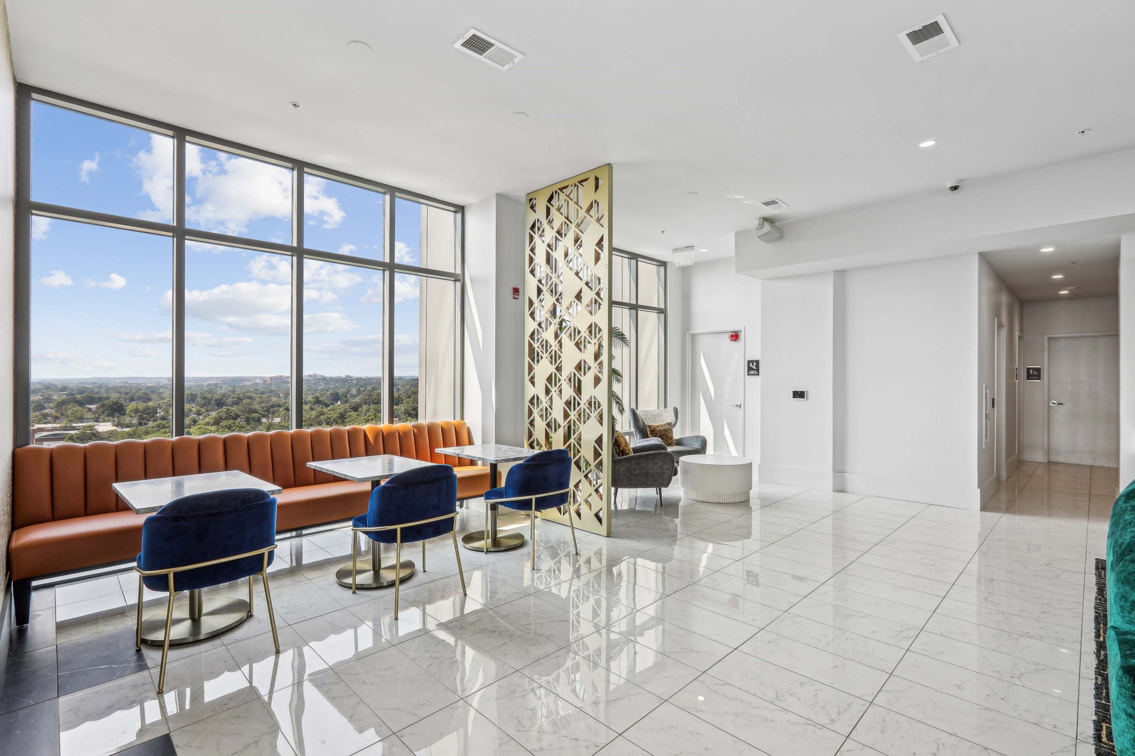 A modern interior space with large windows, featuring seating areas with orange and blue chairs, a decorative partition, and polished marble flooring.