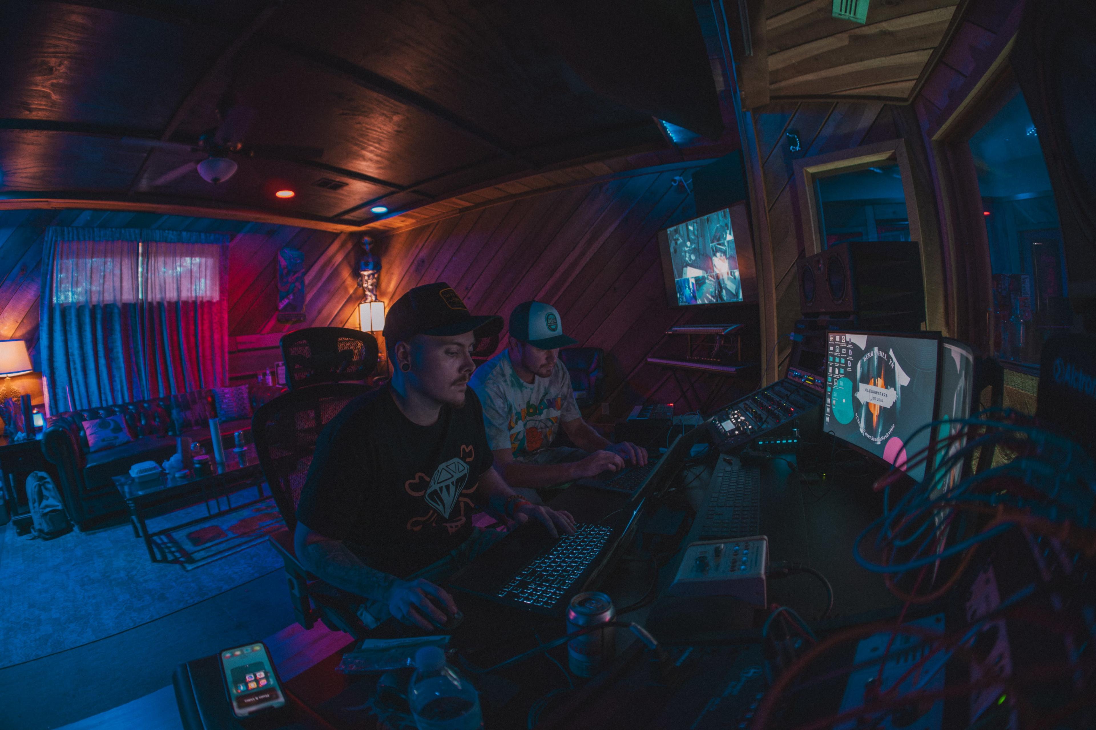 Two individuals are working on computers in a dimly lit recording studio adorned with colorful lighting and wooden paneling.