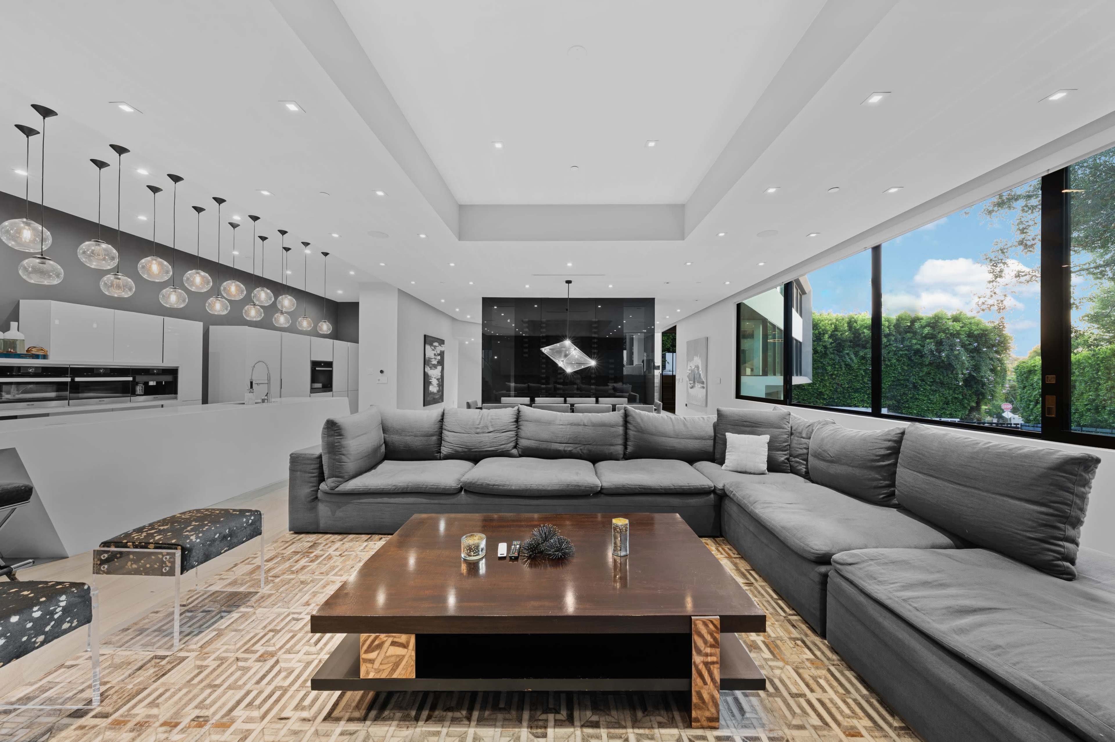 The image shows a spacious modern living room with a large sectional sofa, a wooden coffee table, and large windows allowing natural light to fill the space.