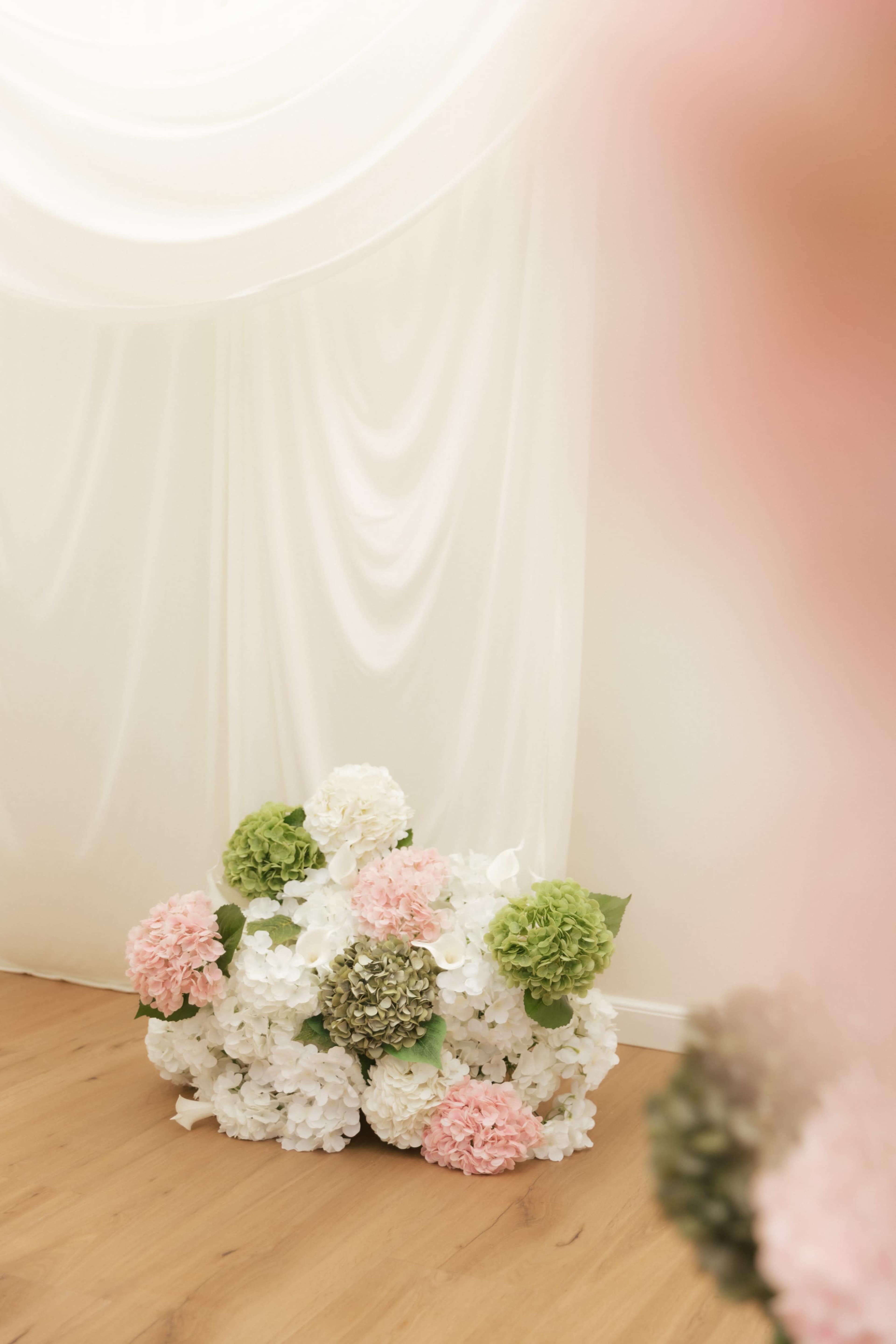 A arrangement of artificial flowers in pink, white, and green is positioned on a wooden floor in front of a white draped backdrop.