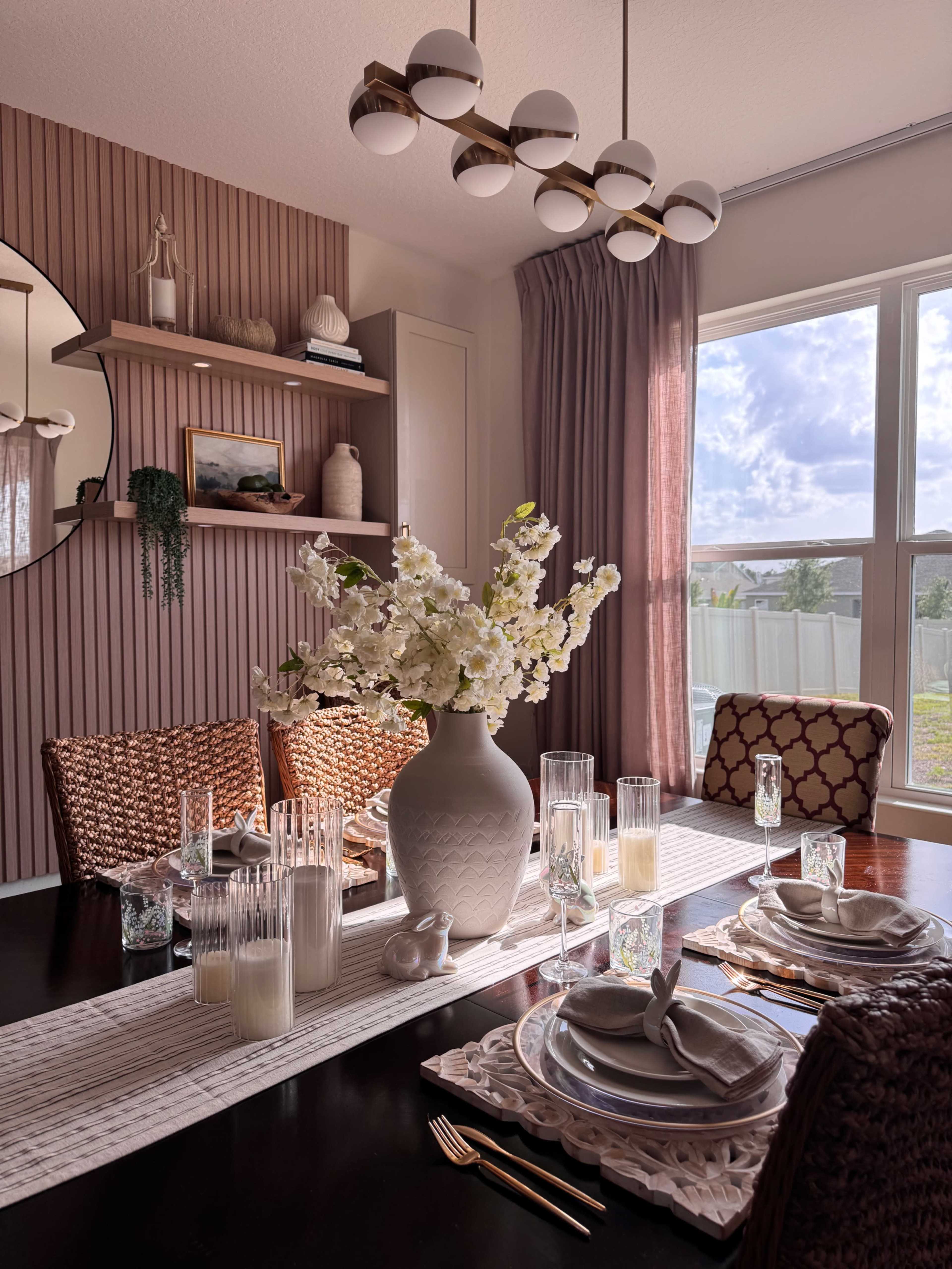 A dining room features a dark wooden table set with elegant tableware and a large vase of flowers, surrounded by woven chairs and illuminated by a modern light fixture.