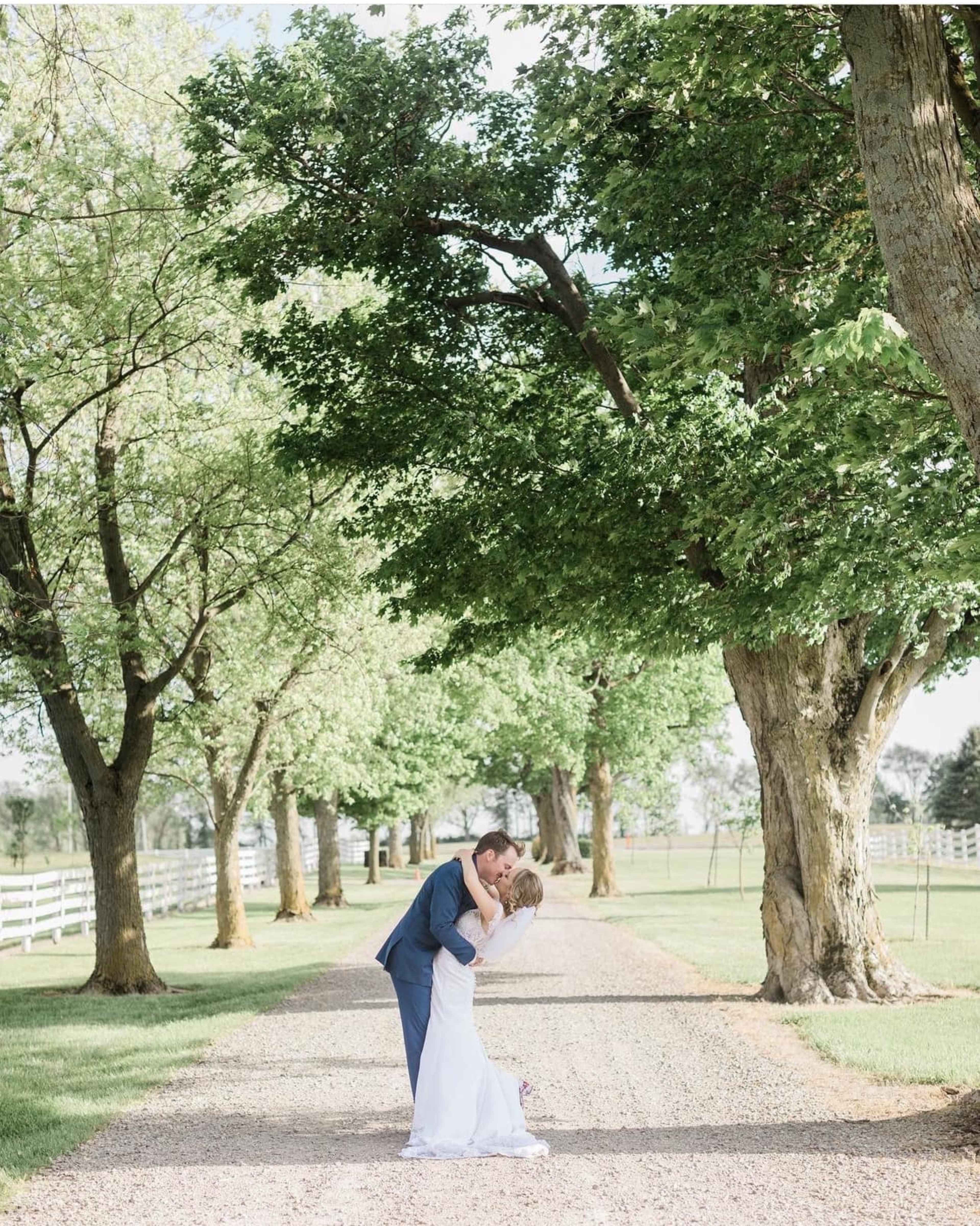 A couple embraces on a gravel path lined with trees and a white fence.
