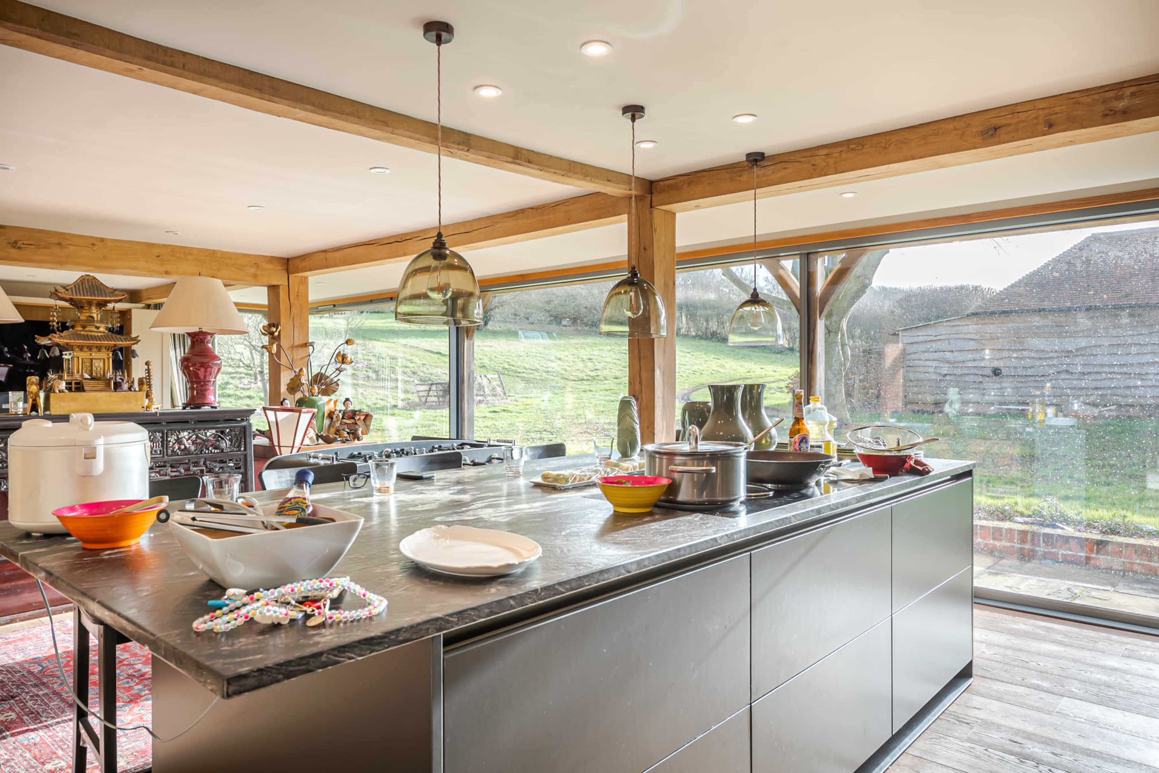 A modern kitchen with a large island, scattered dishes, and large windows overlooking a green landscape.