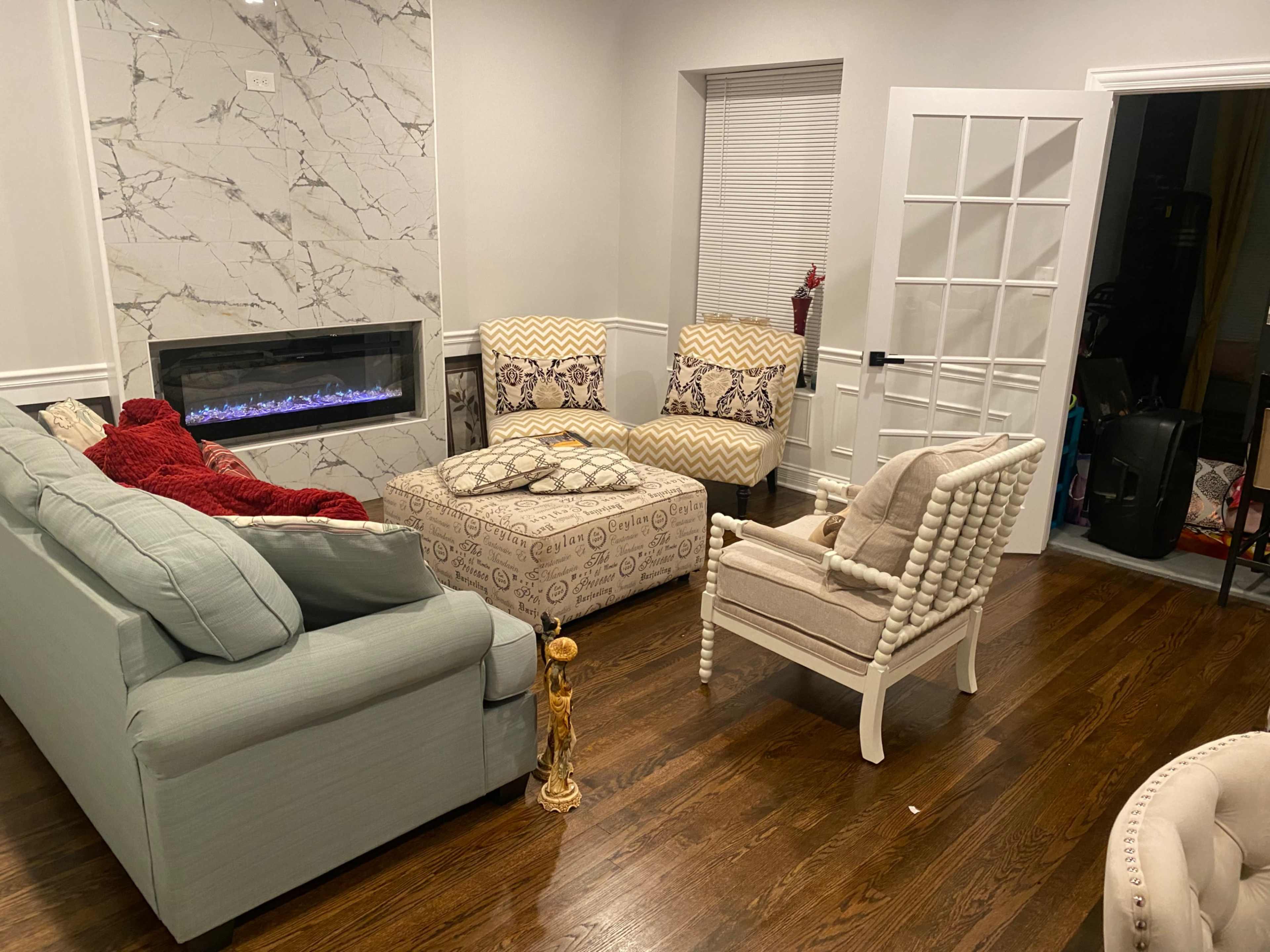 The living room features a set of light-colored furniture arranged around a modern electric fireplace, with a decorative gold figure in the foreground.