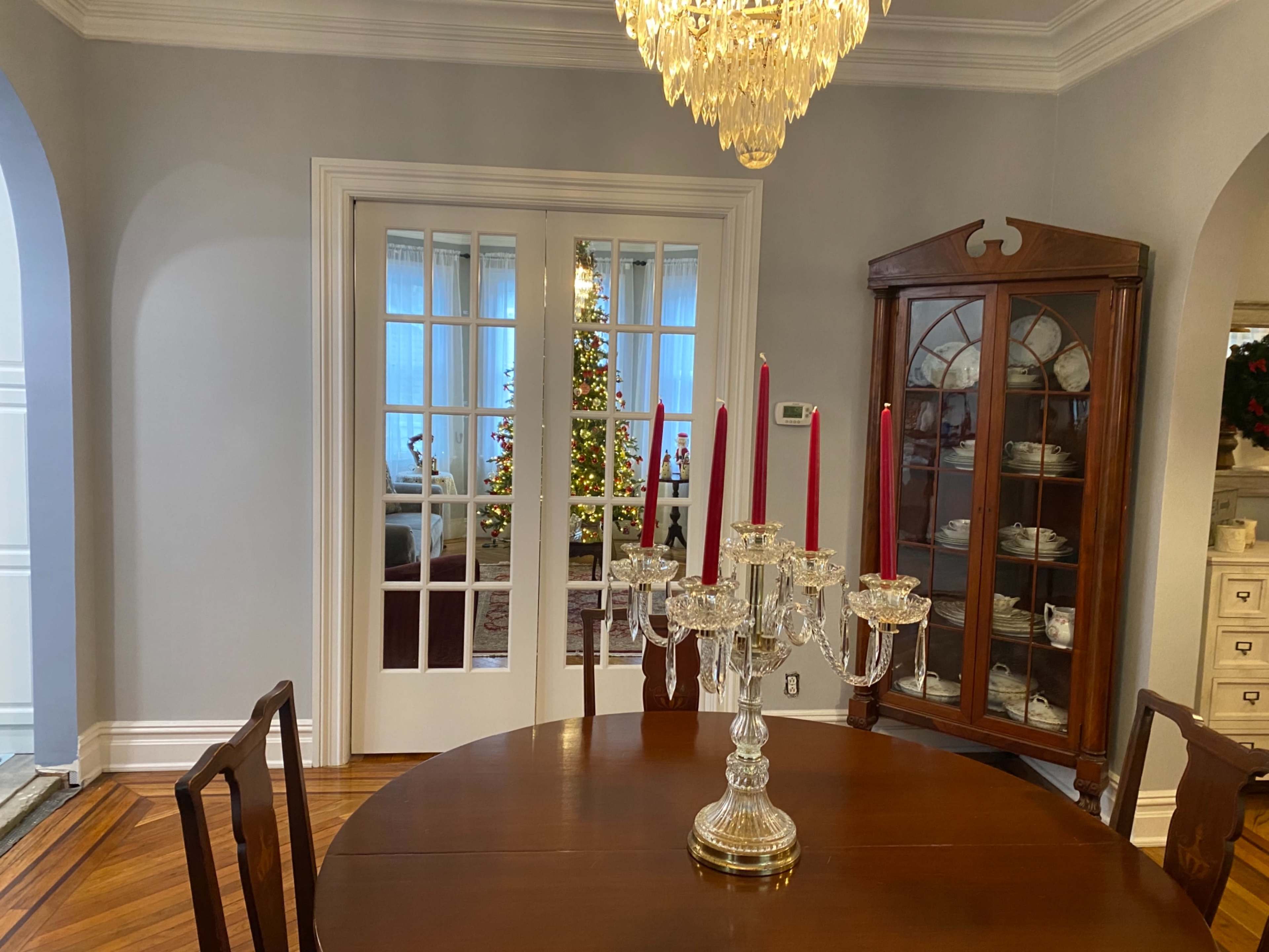 A dining room features a wooden table with a crystal candleholder and red candles, while a glass door reveals a glimpse of a Christmas tree beyond.