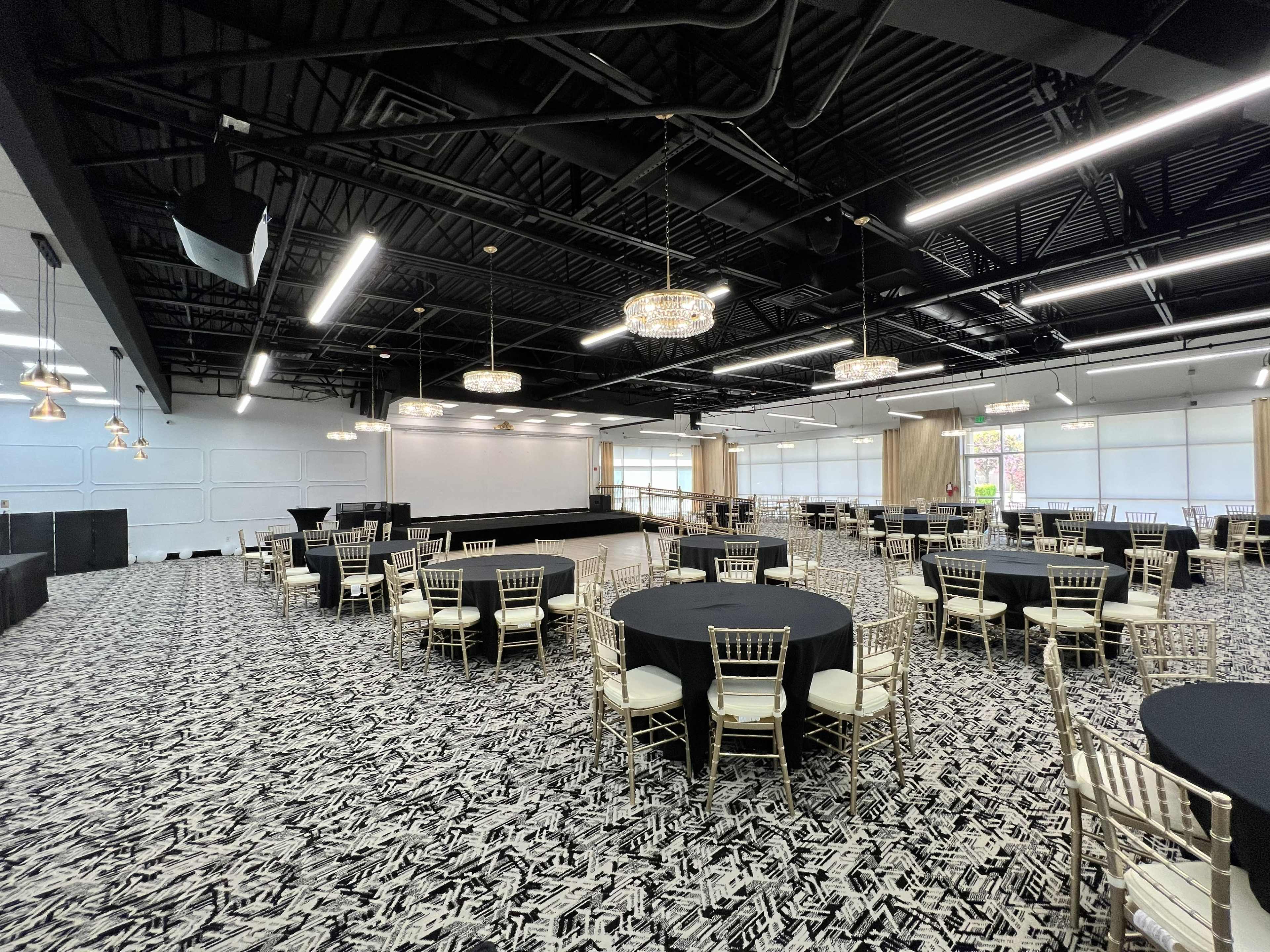 The image shows an event space with several round tables covered in black linens, gold chairs, and a stage area at the front, illuminated by overhead lighting.