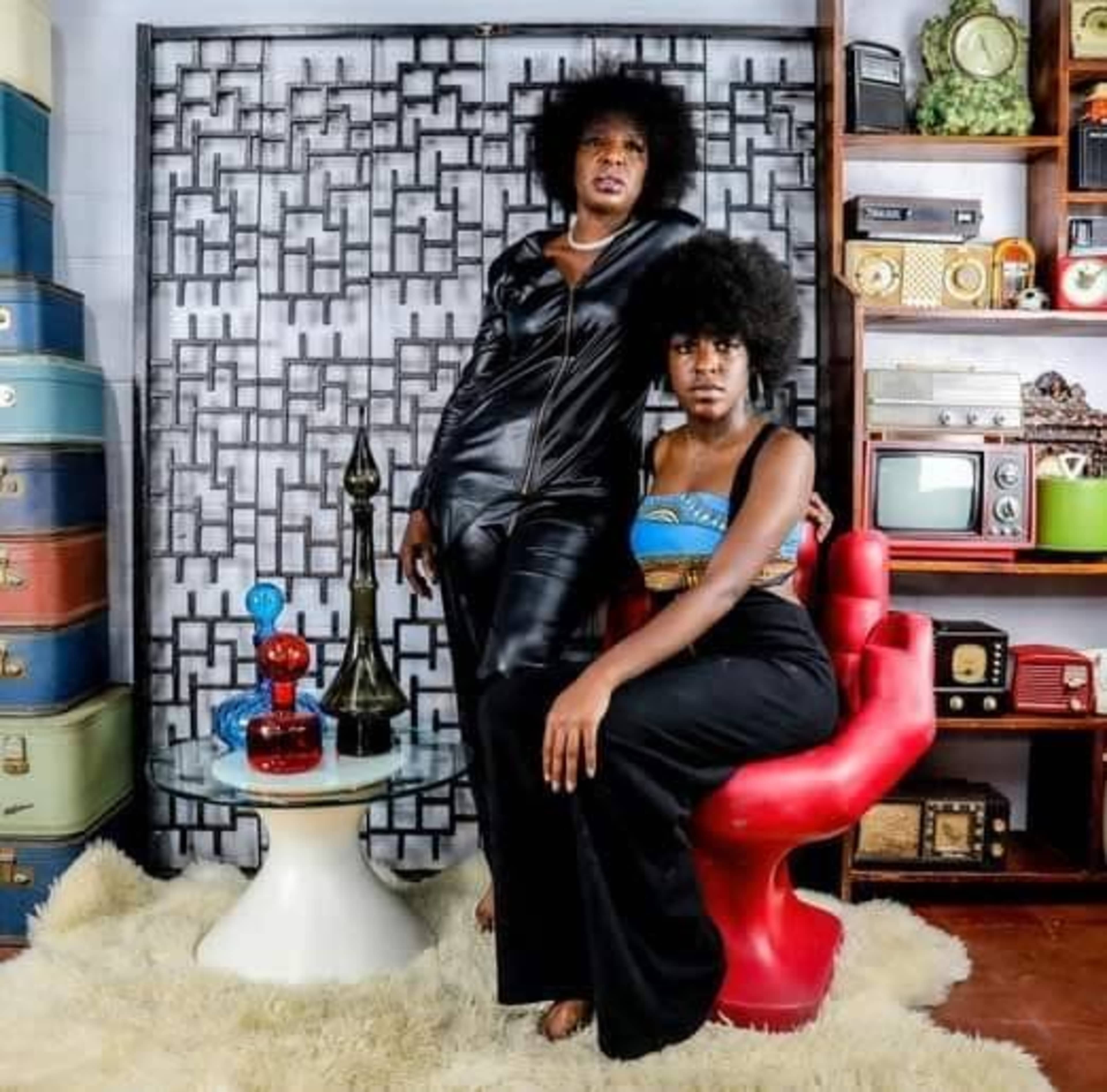 Two women with afro hairstyles pose in a stylish room featuring a decorative wall, a vintage-style shelf, and colorful accessories.