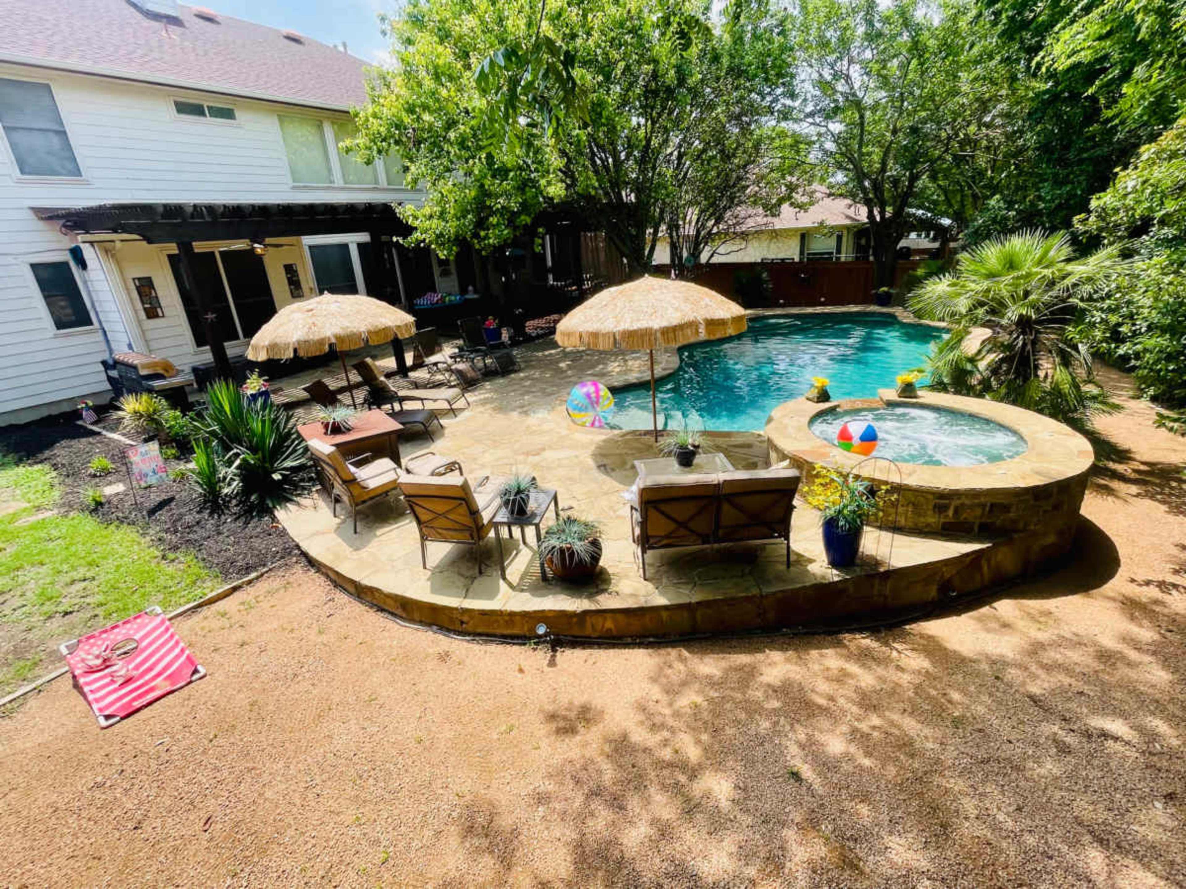 The image shows a backyard pool area with lounge chairs, two umbrellas, and a hot tub surrounded by greenery and stone pathways.