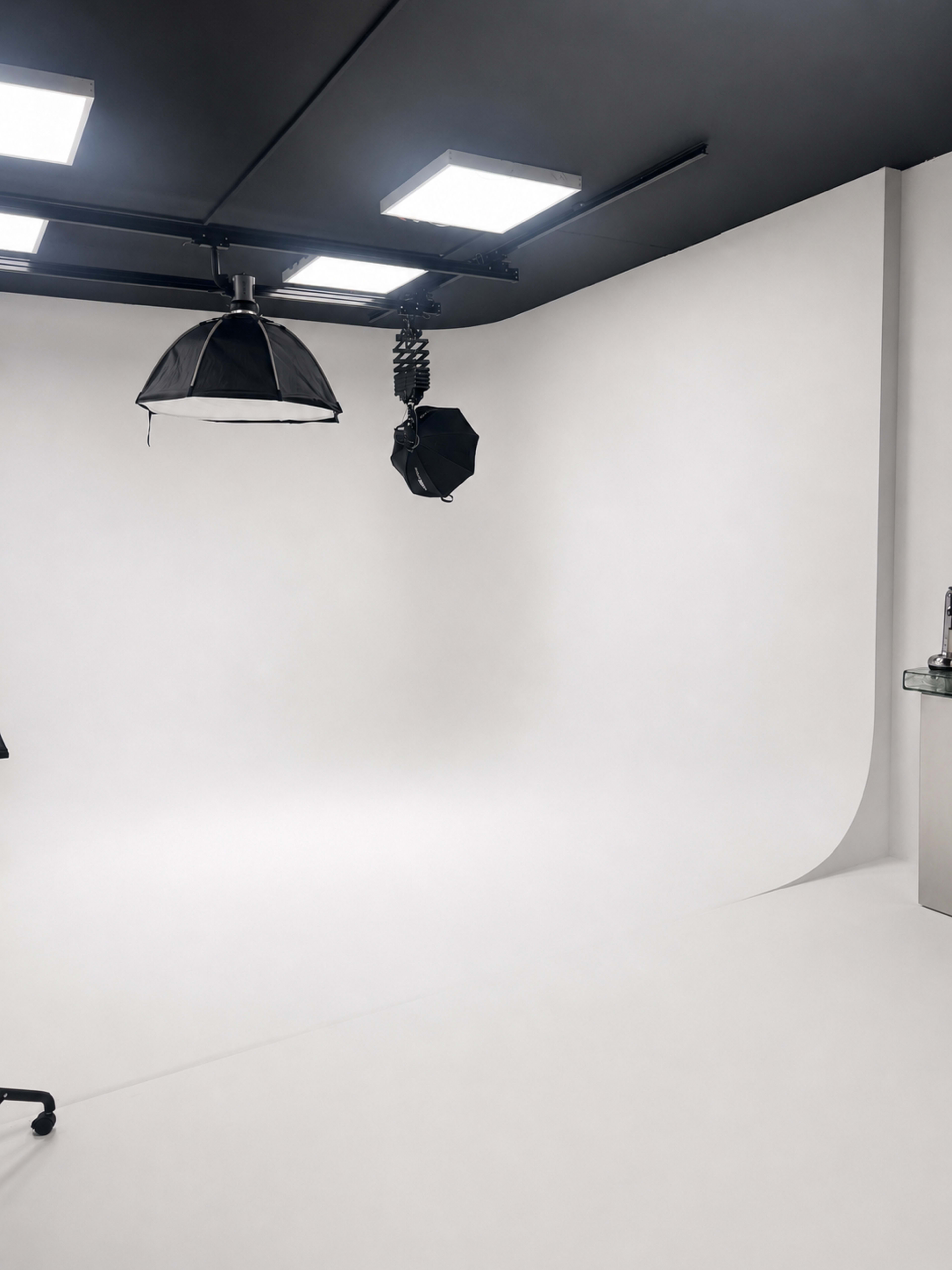 The image shows a minimalist photography studio with a seamless white backdrop, softbox lighting, and a small table in the corner.
