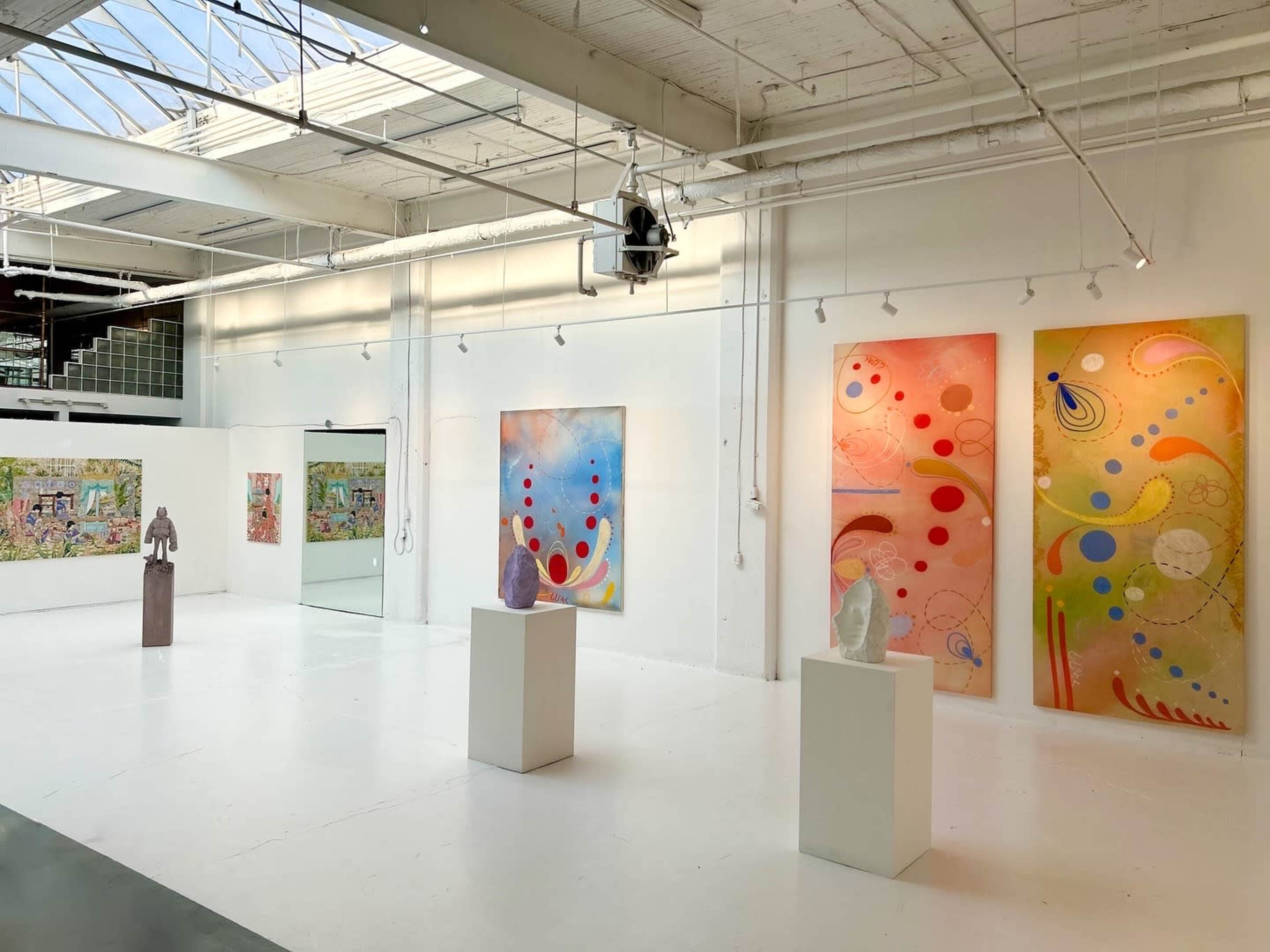 The image shows an art gallery with colorful abstract paintings on the walls and several sculptures displayed on pedestals in a well-lit space.