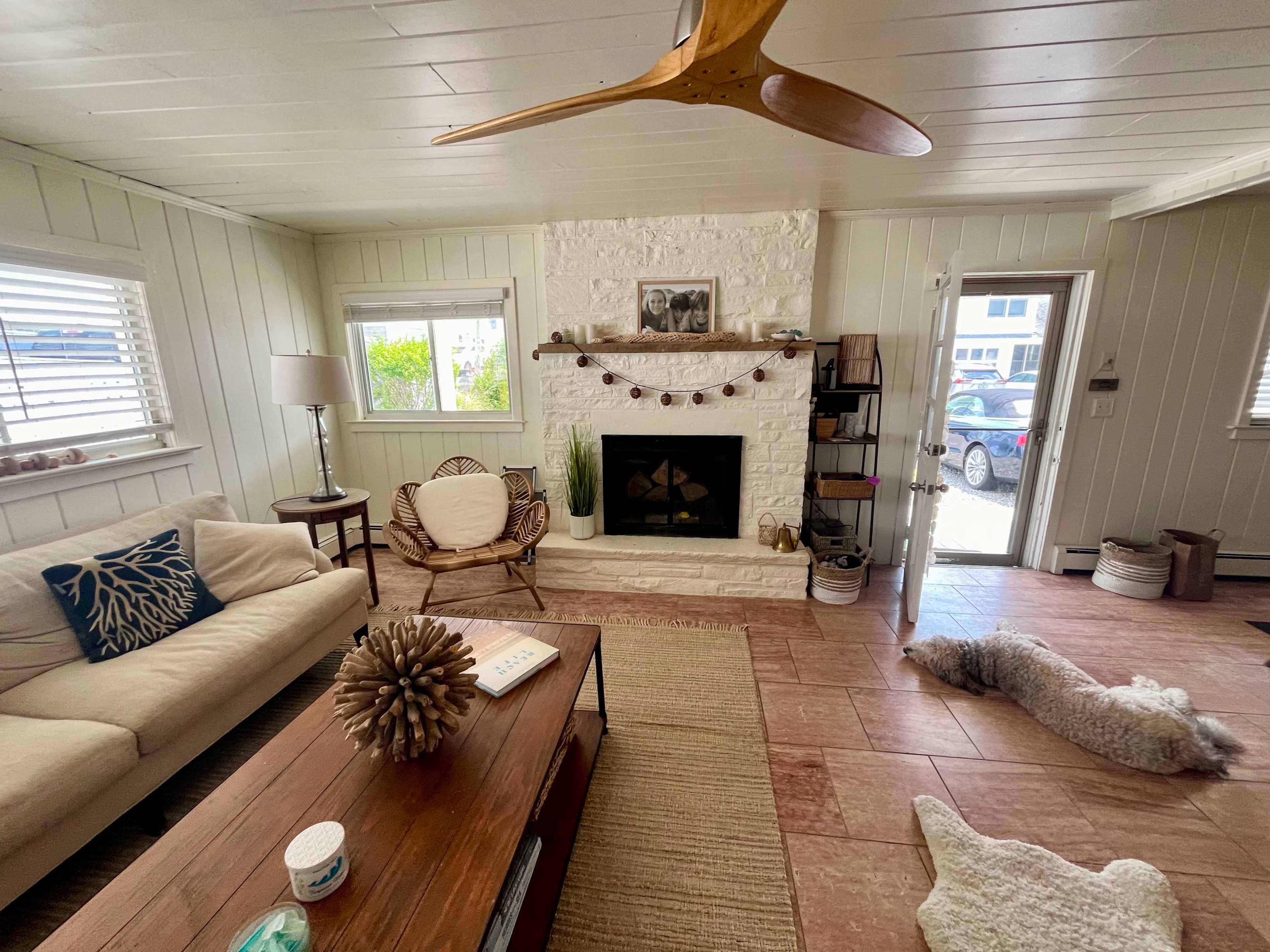 A cozy living room with a white stone fireplace, a wooden coffee table, a sofa, and a door leading outside, while a dog lies on the floor.