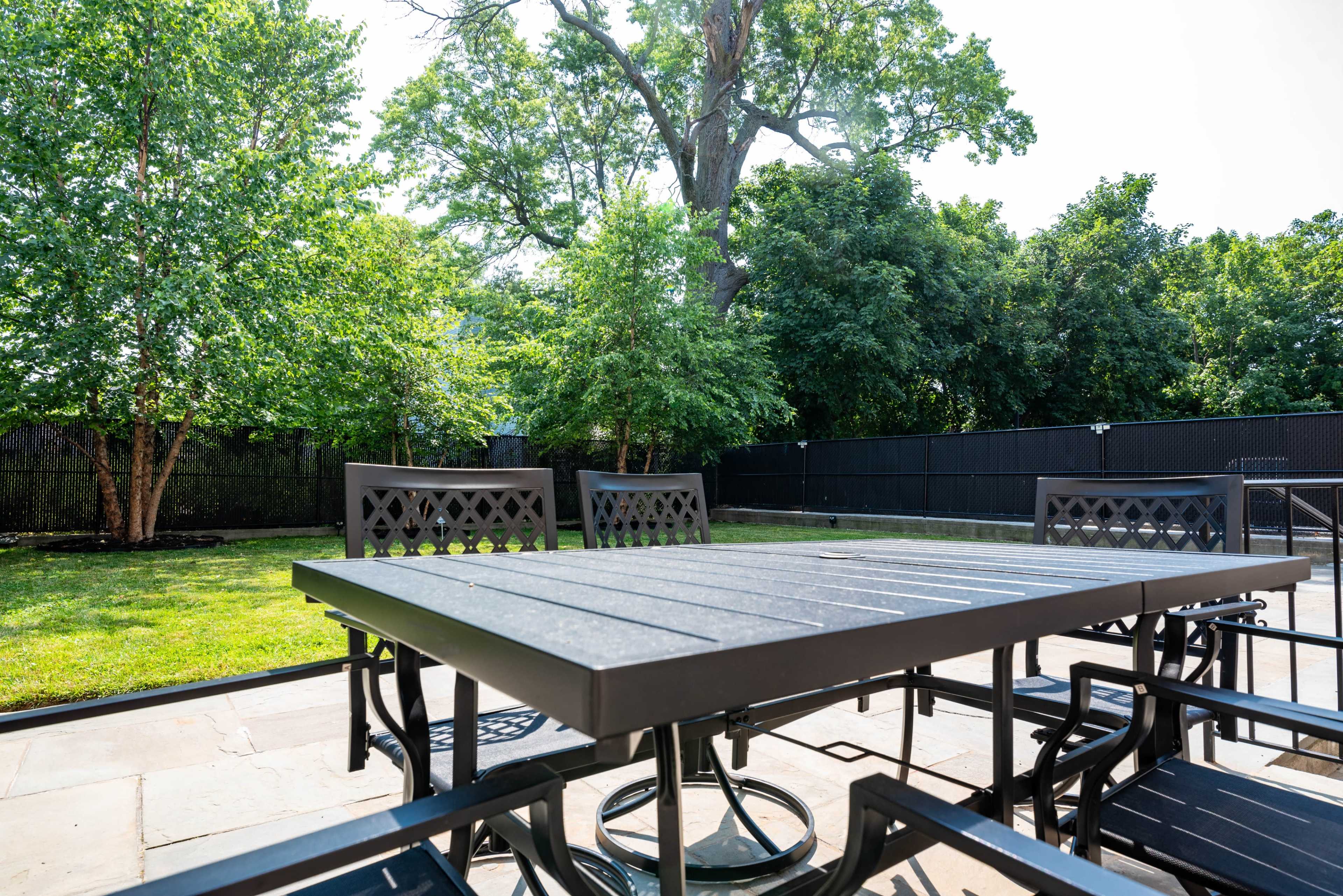 The image shows a patio area with a metal table surrounded by chairs, set against a backdrop of green trees and a black fence.