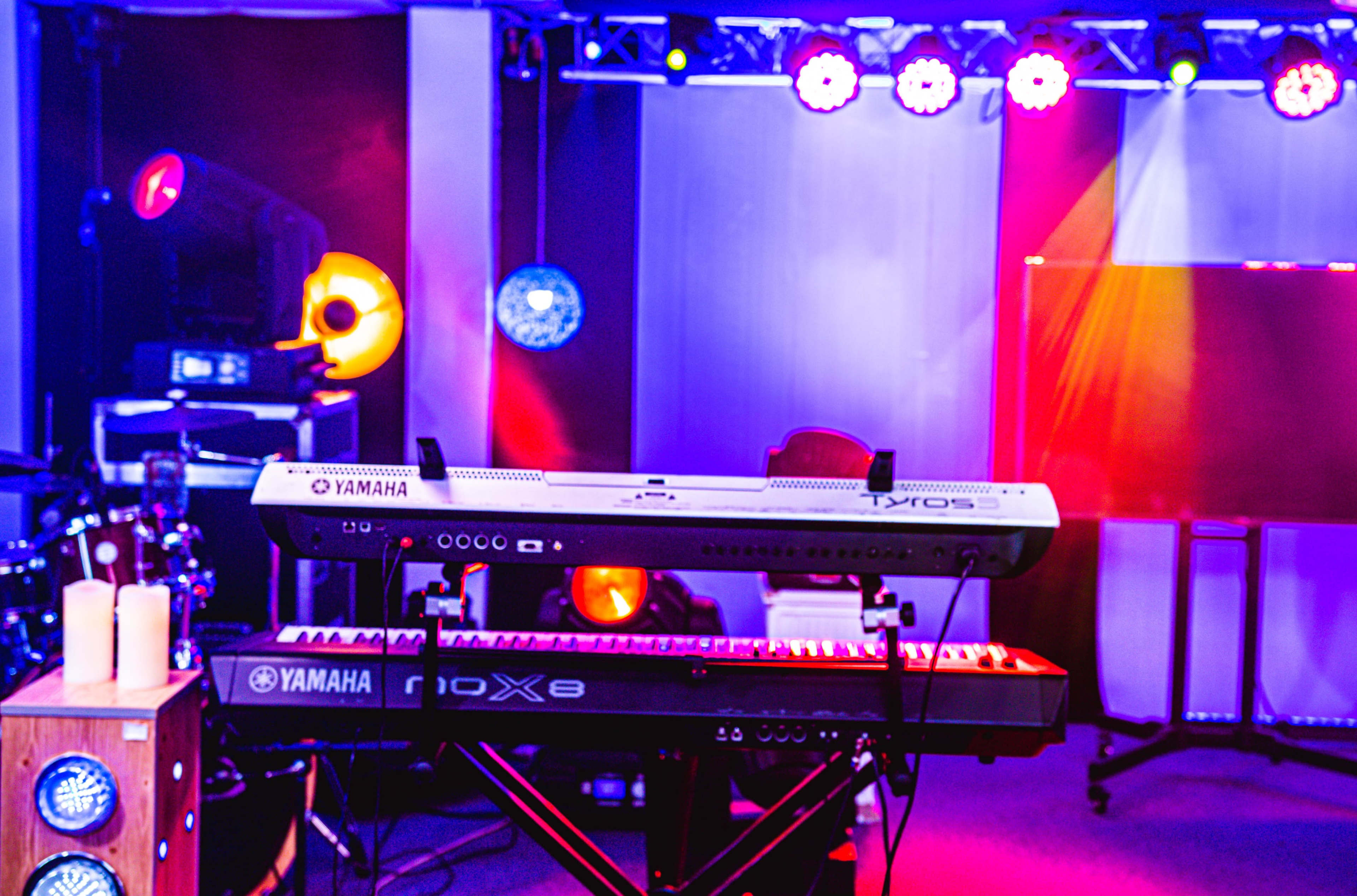 A stage setup featuring two Yamaha keyboards, a drum set, and colorful stage lights illuminating the area.