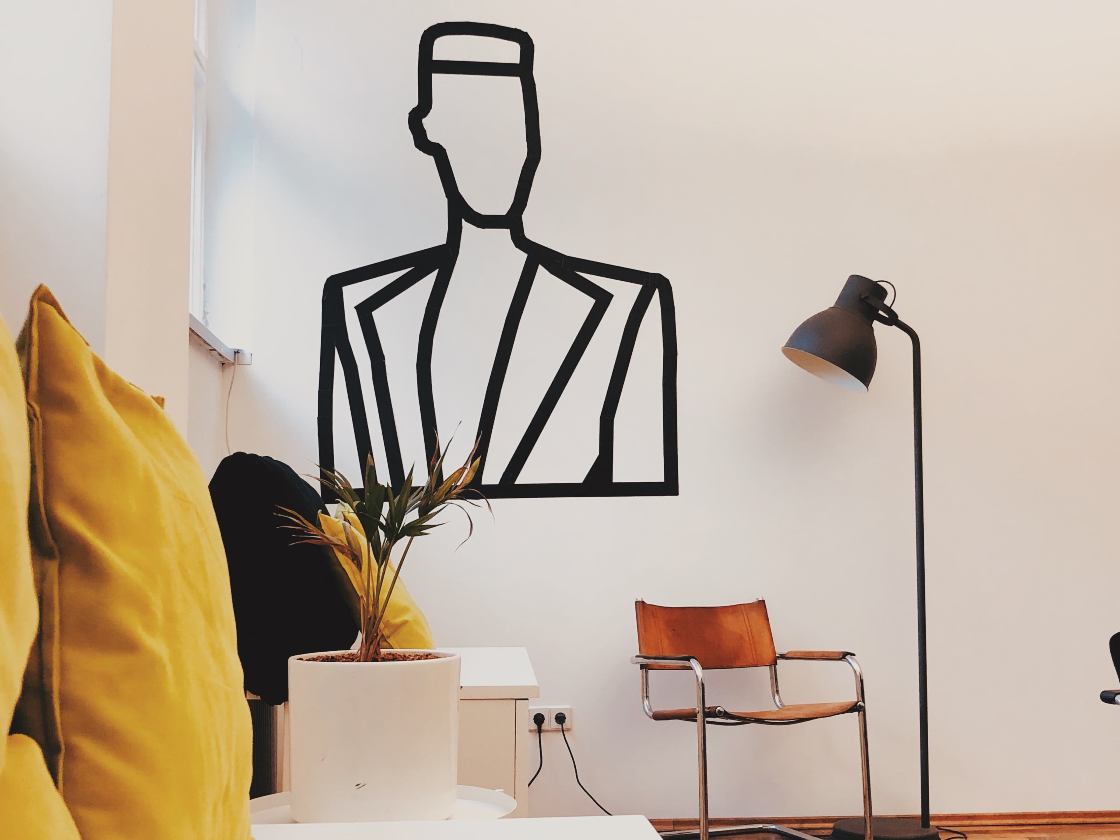 A modern indoor space with a minimalist wall mural of a suit figure, a yellow cushion on a sofa, a potted plant, and a wooden chair beside a standing lamp.