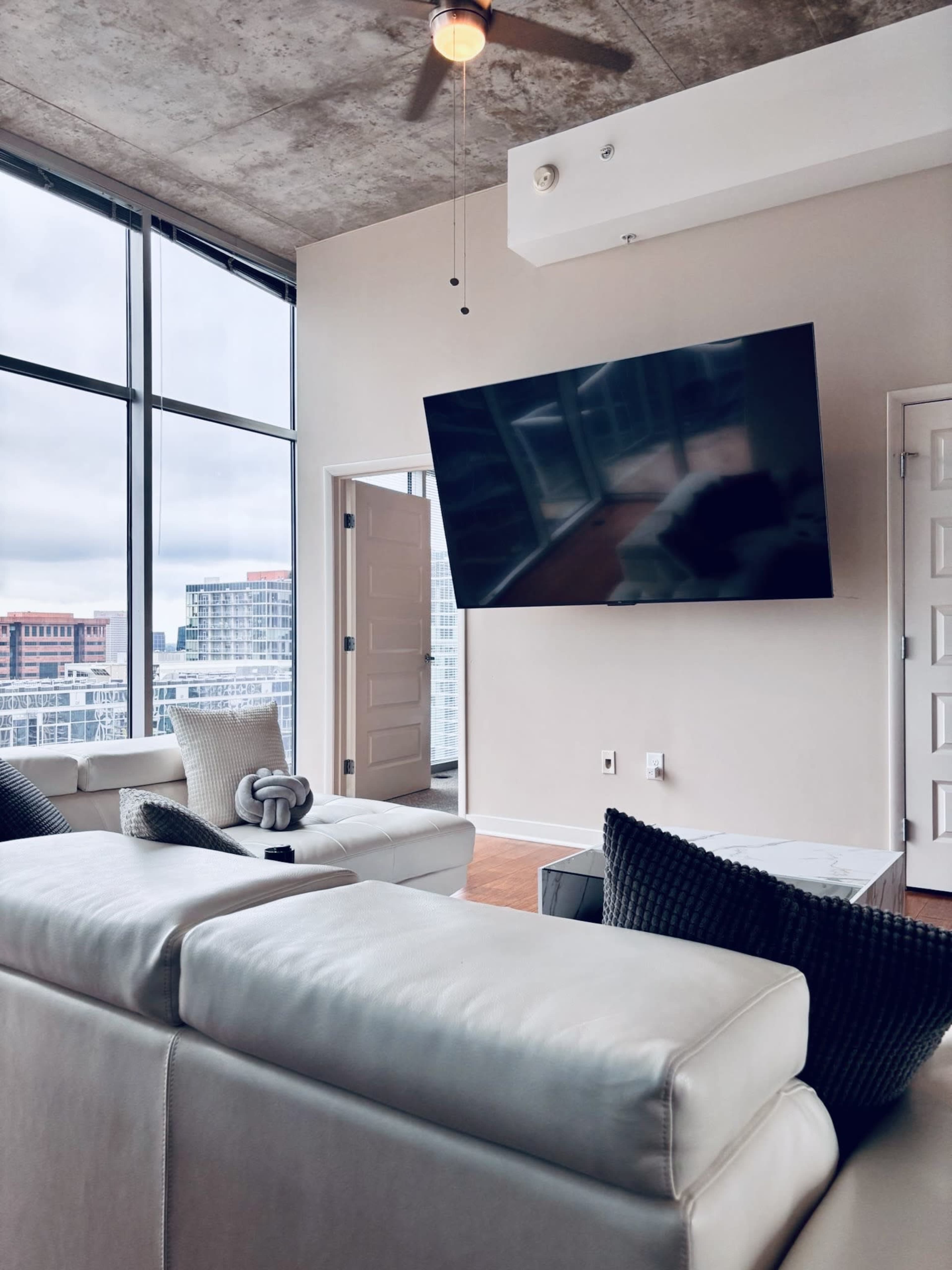 The image shows a modern living room with large windows, a wall-mounted television, and a light-colored leather sofa.