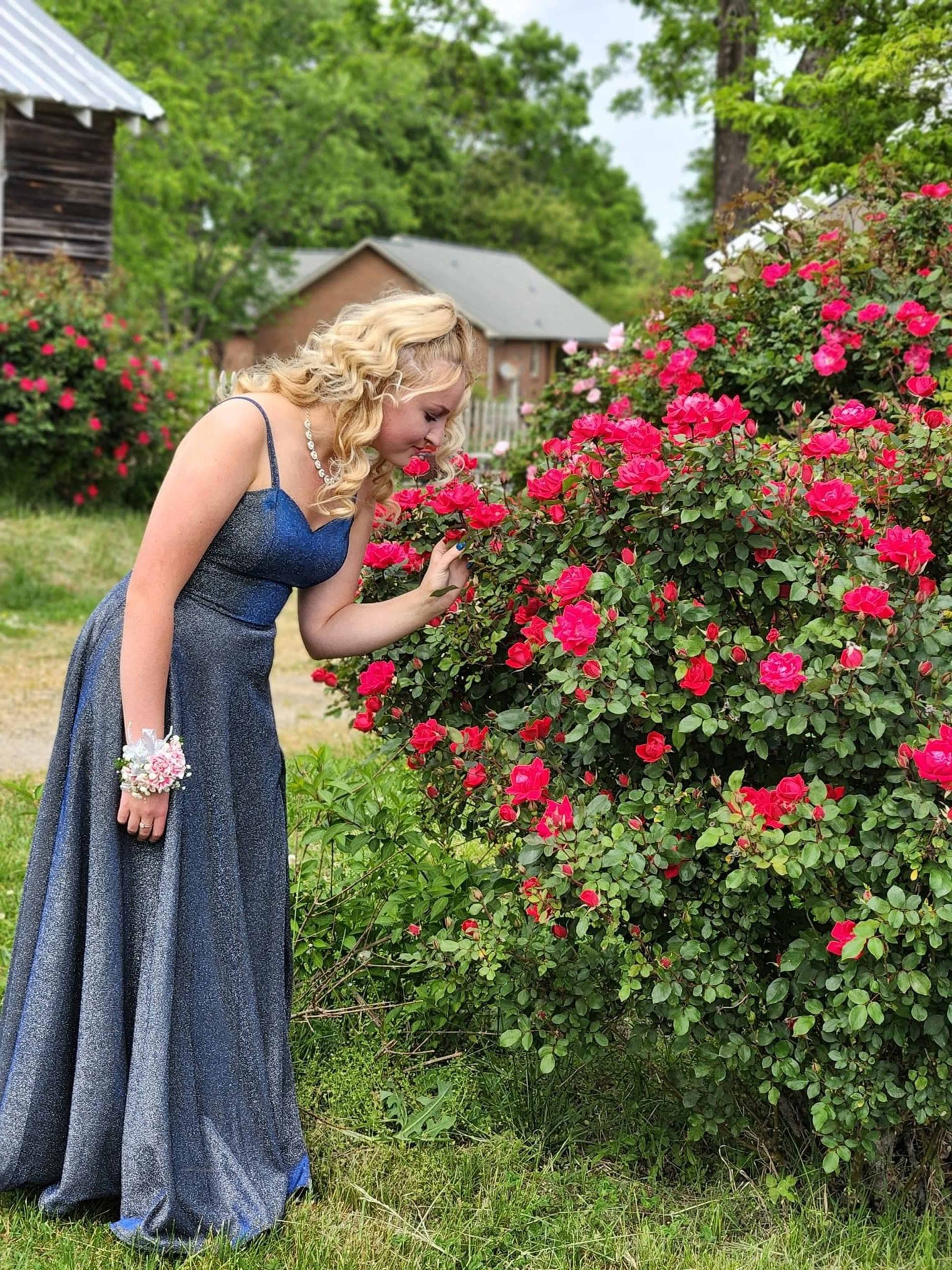 A young woman in a formal dress stands beside a flowering rose bush, admiring the vibrant pink blooms.