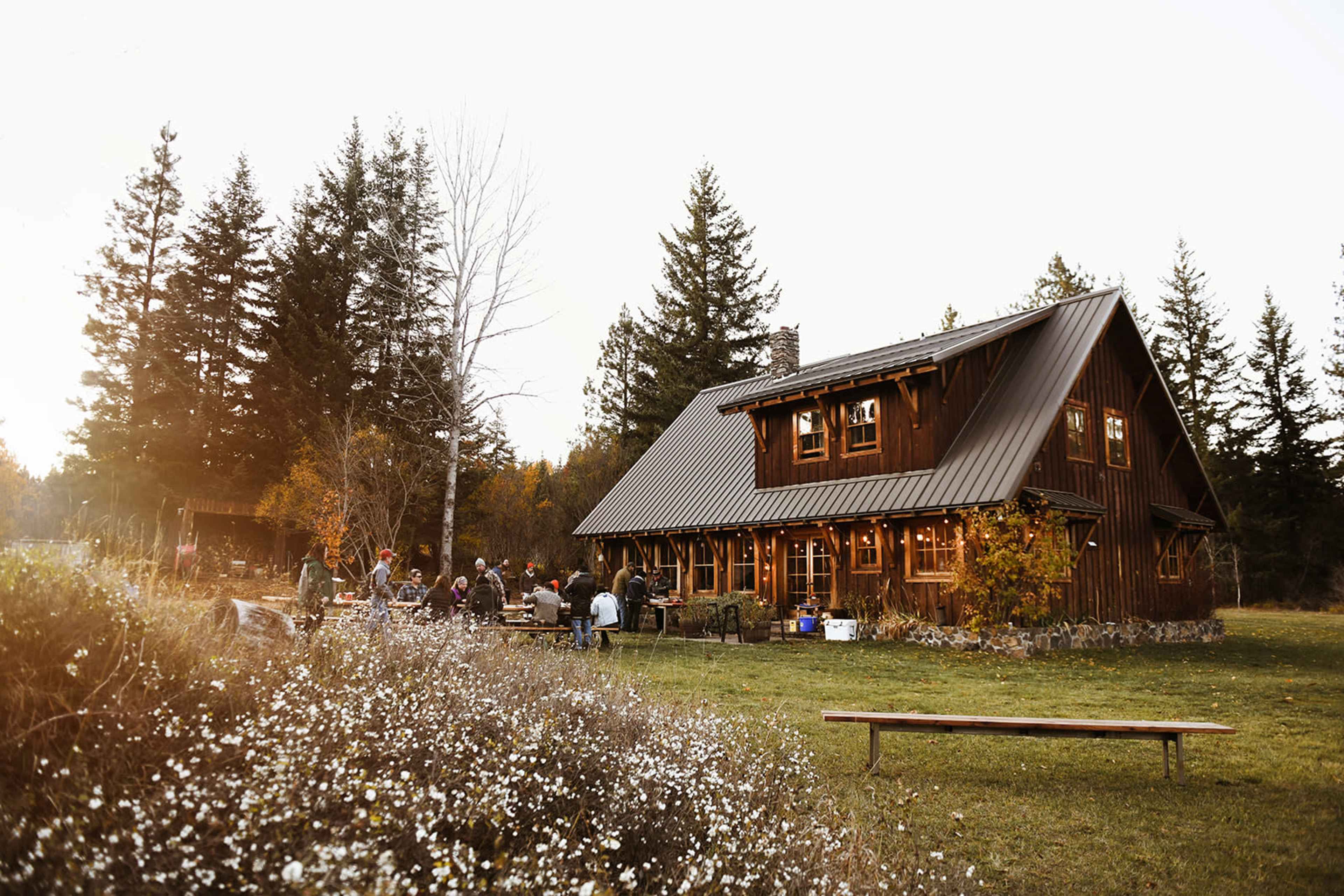 A rustic wooden cabin sits amidst a group of people gathered outside in a natural setting surrounded by trees and wildflowers.