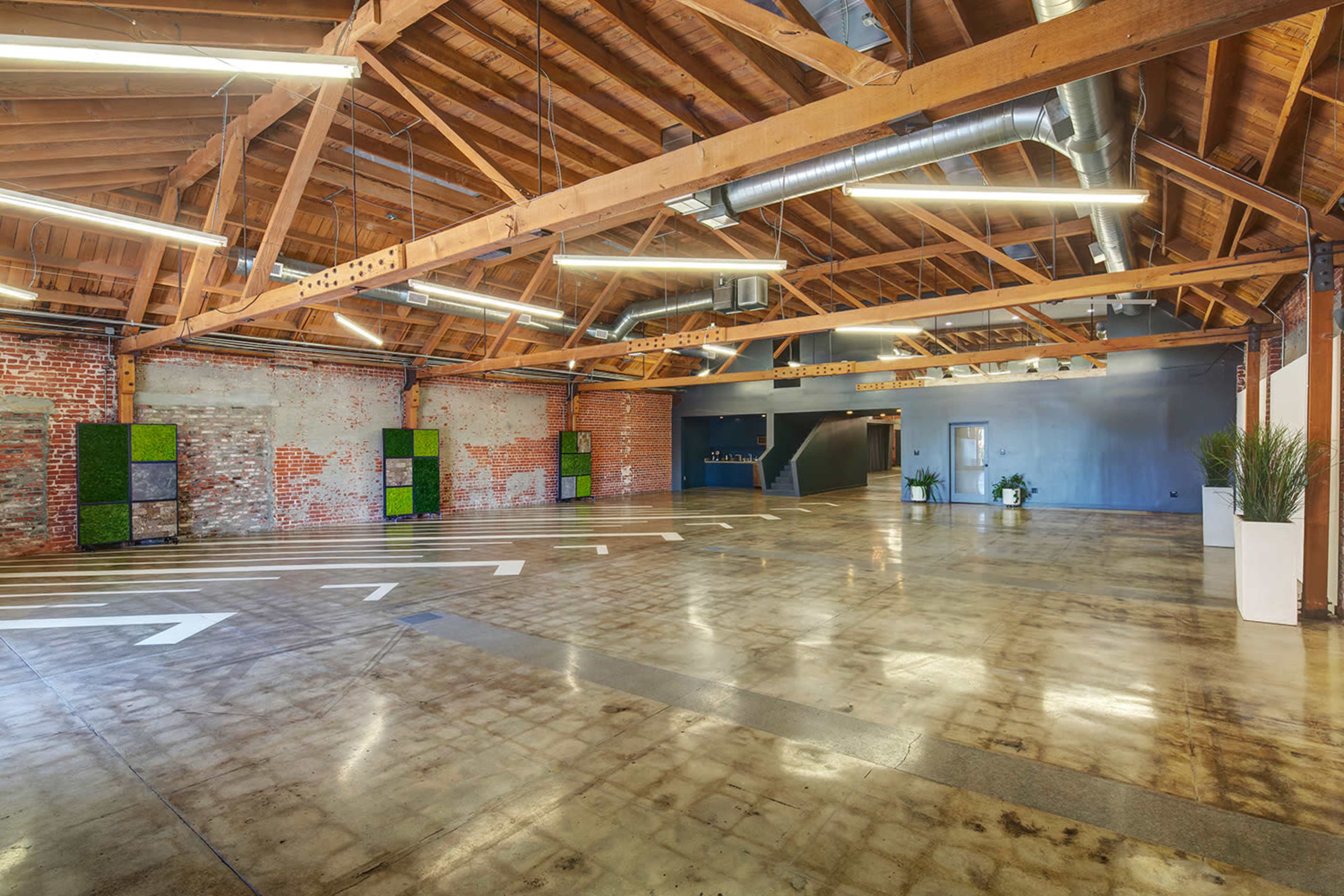 The image shows a spacious industrial-style room with exposed wooden beams, polished concrete flooring, and brick walls, featuring several decorative plants along the edges.