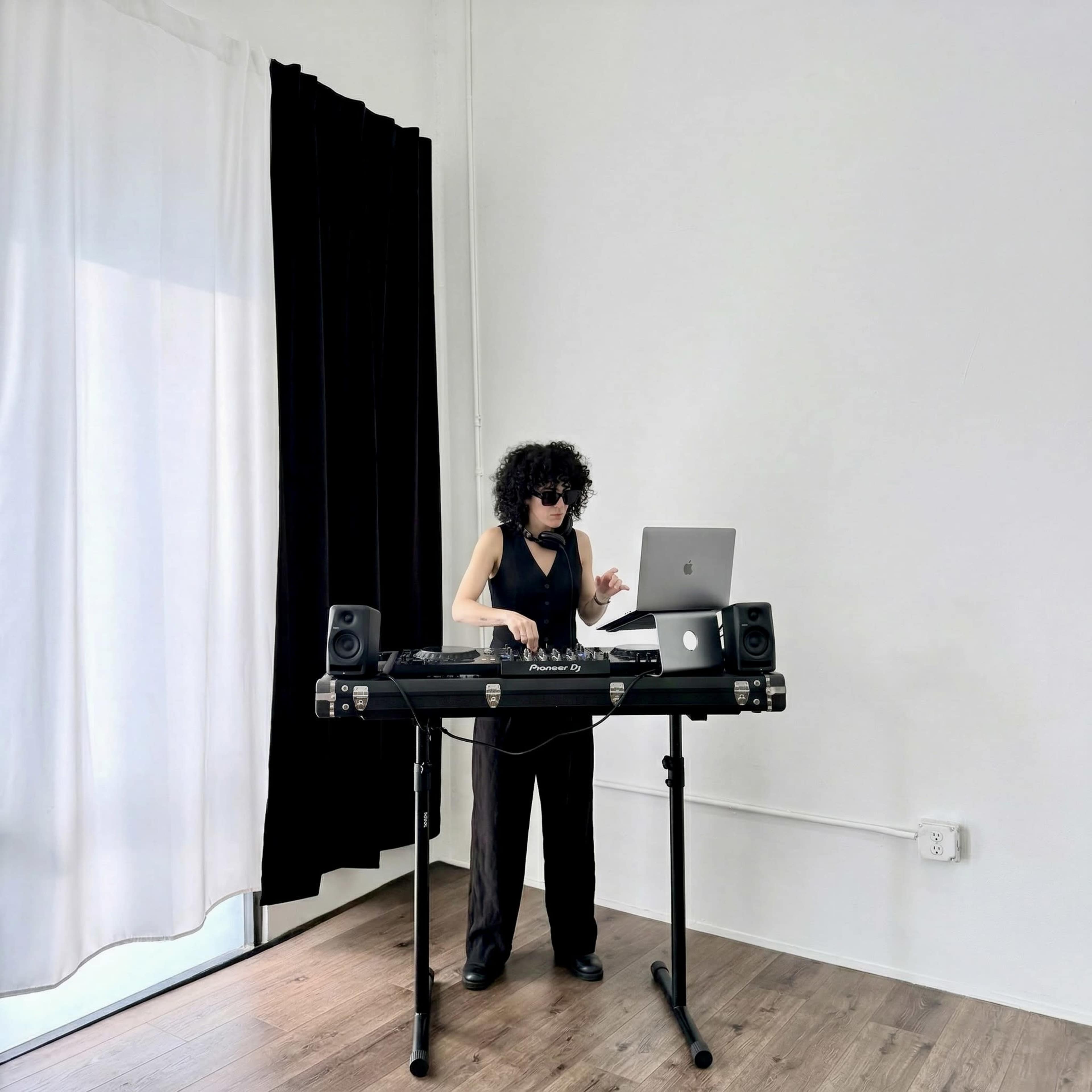 A person with curly hair and sunglasses is mixing music on a DJ setup in a minimalist room with black and white curtains.
