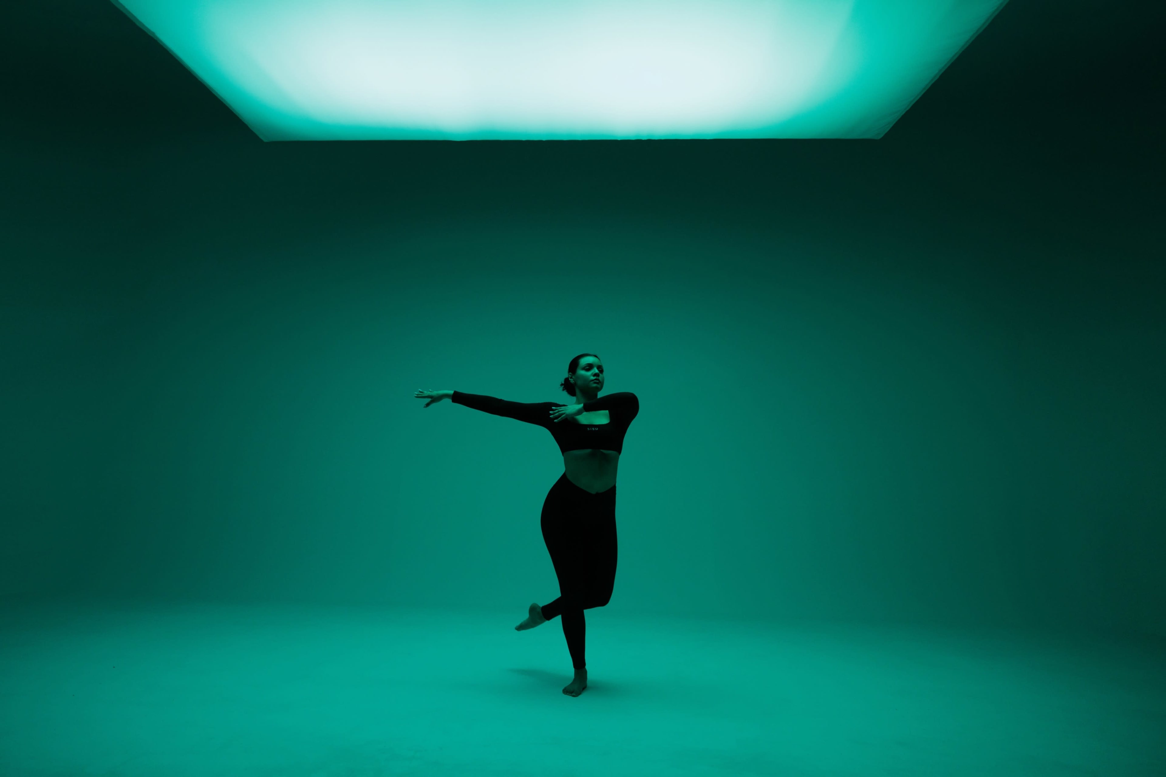 A dancer performs gracefully in a dimly lit studio with an illuminated ceiling casting a green hue.
