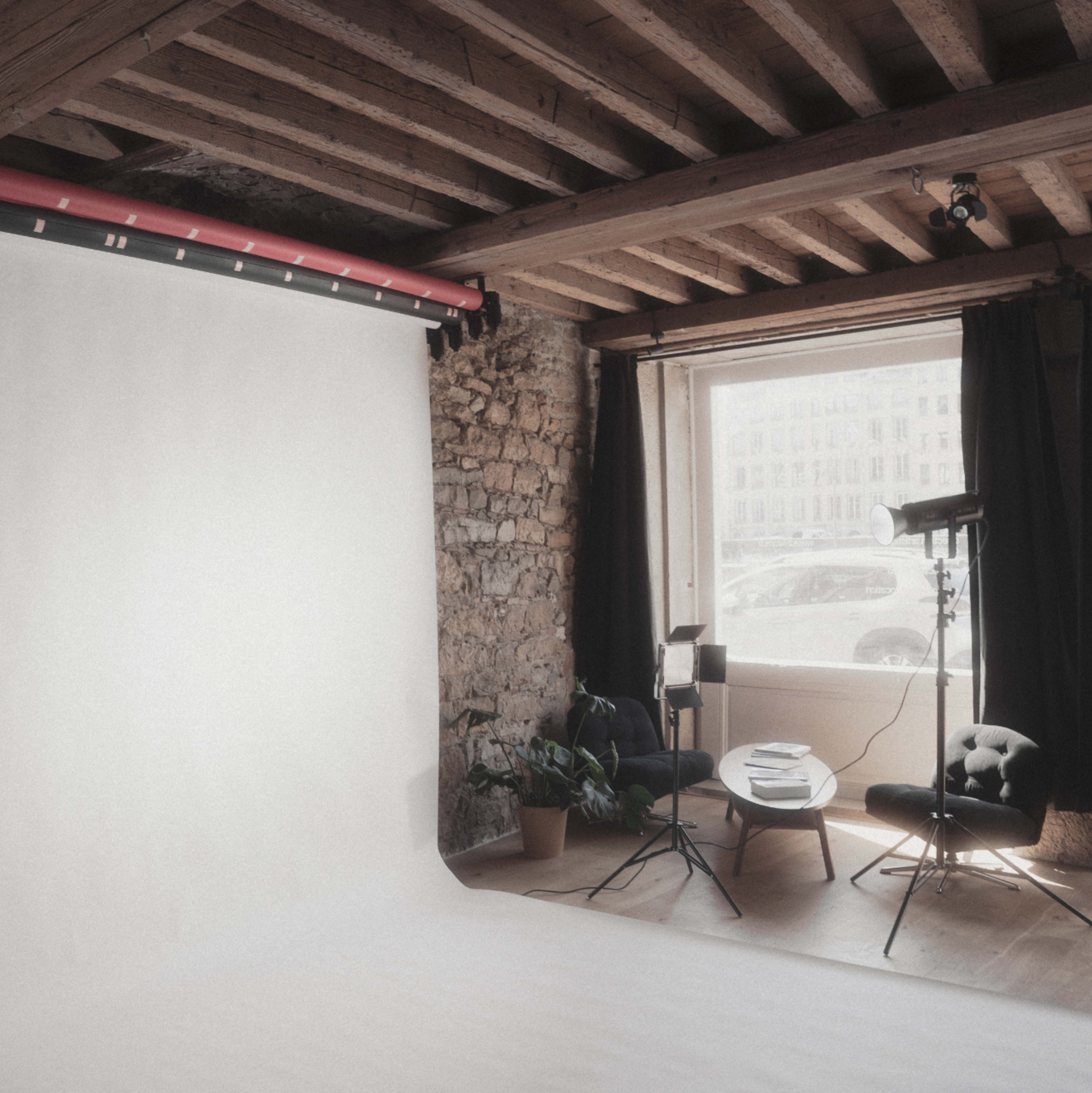 The image depicts a photography studio with a seamless white backdrop, soft natural light coming through a window, and minimal furniture including a chair and potted plant.