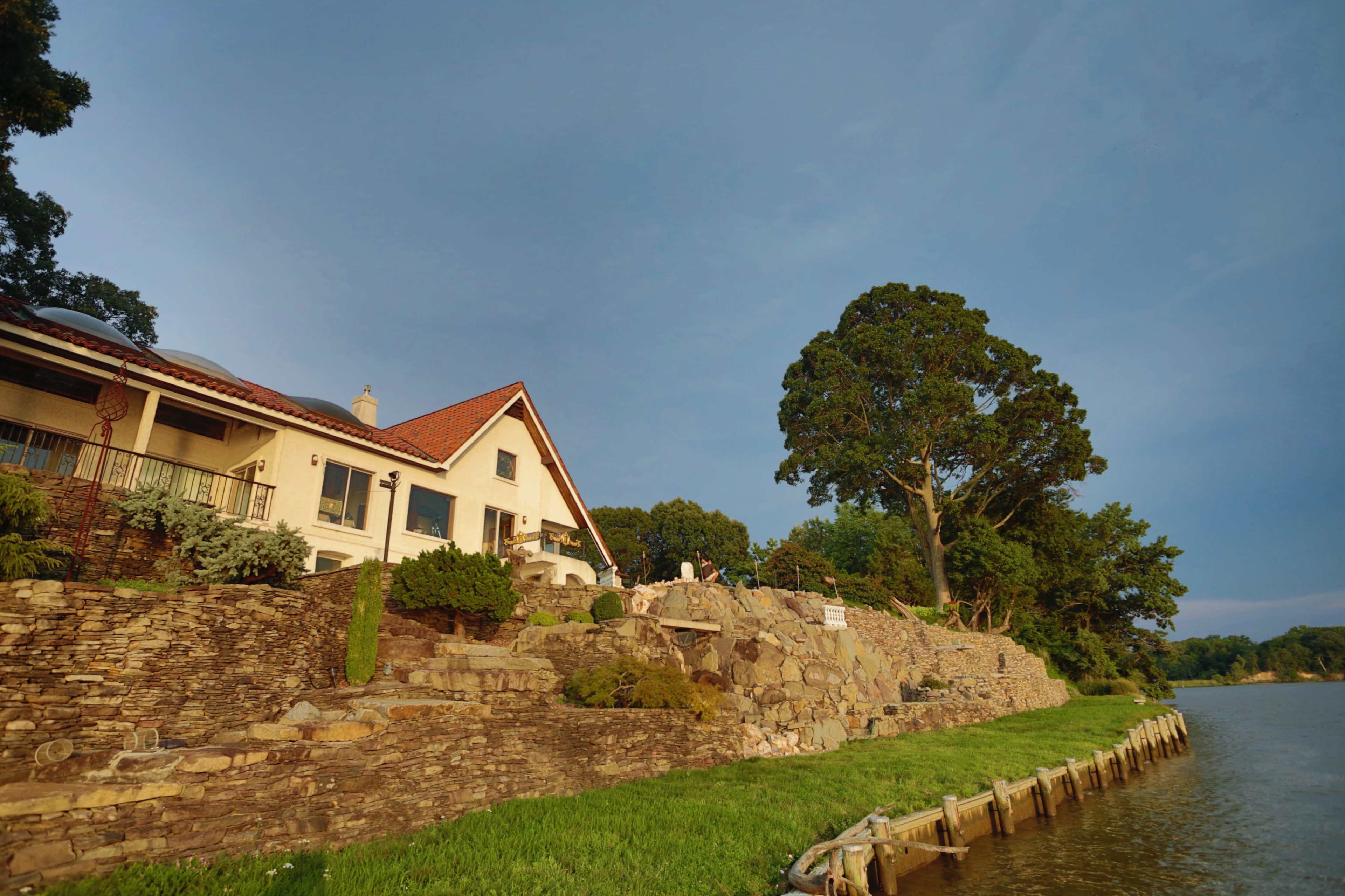 A large stone house with a red-tiled roof overlooks a calm river, surrounded by trees and grass.