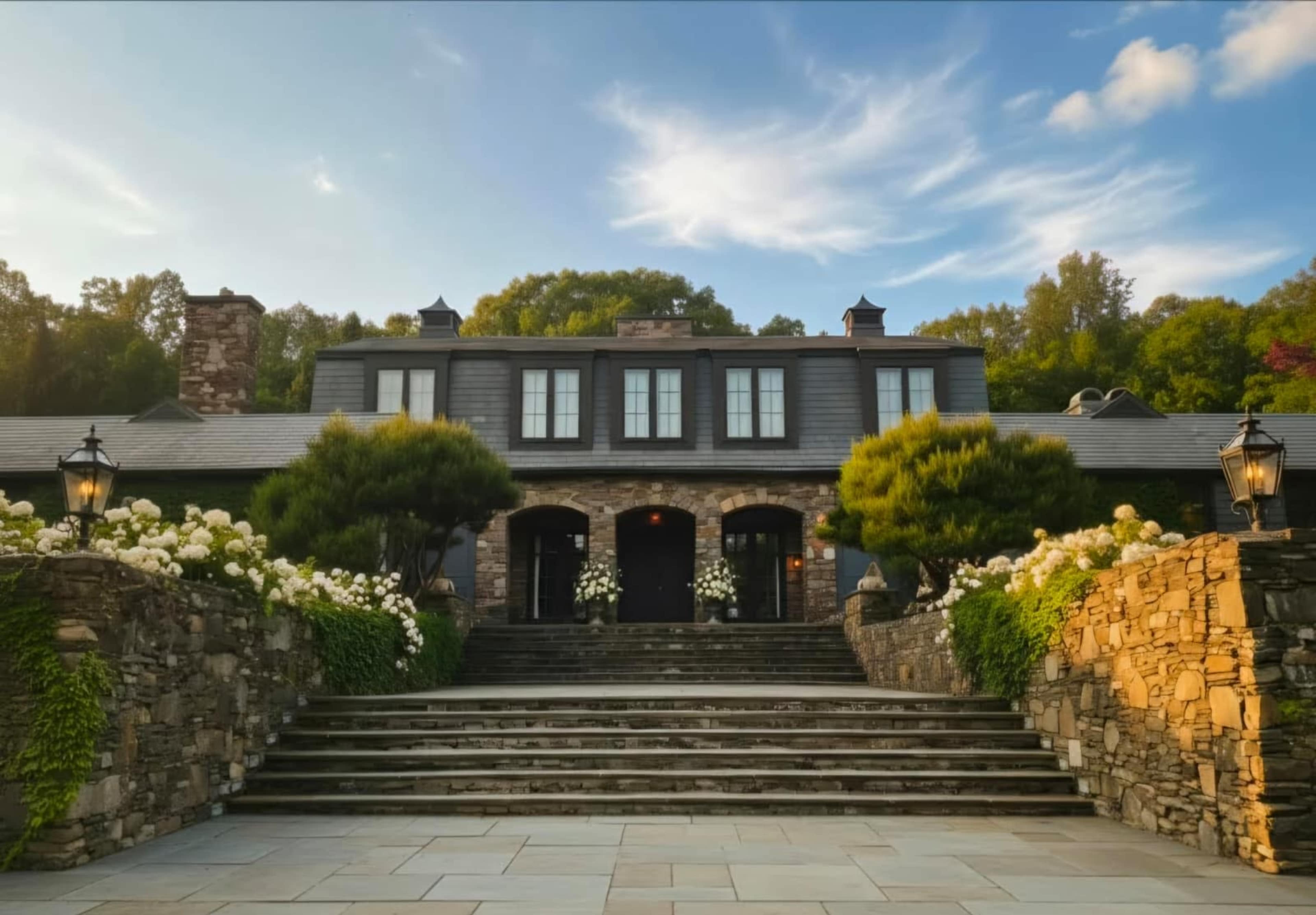 The image shows a large stone mansion with lush landscaping, featuring a set of stairs leading to the entrance, flanked by lanterns and shrubs.