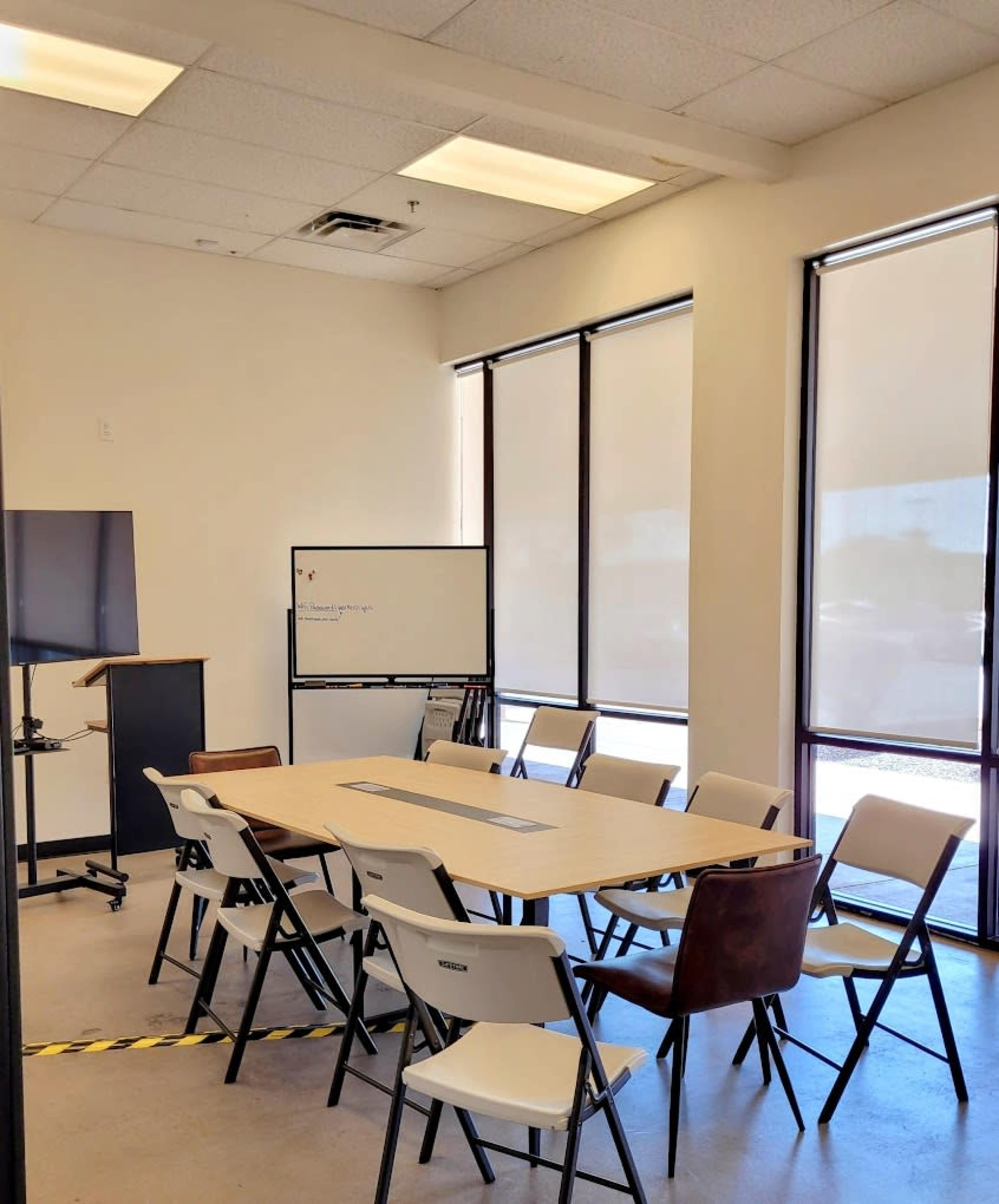 A conference room features a large table surrounded by chairs, a whiteboard, and a television on a stand, with natural light filtering through large windows.