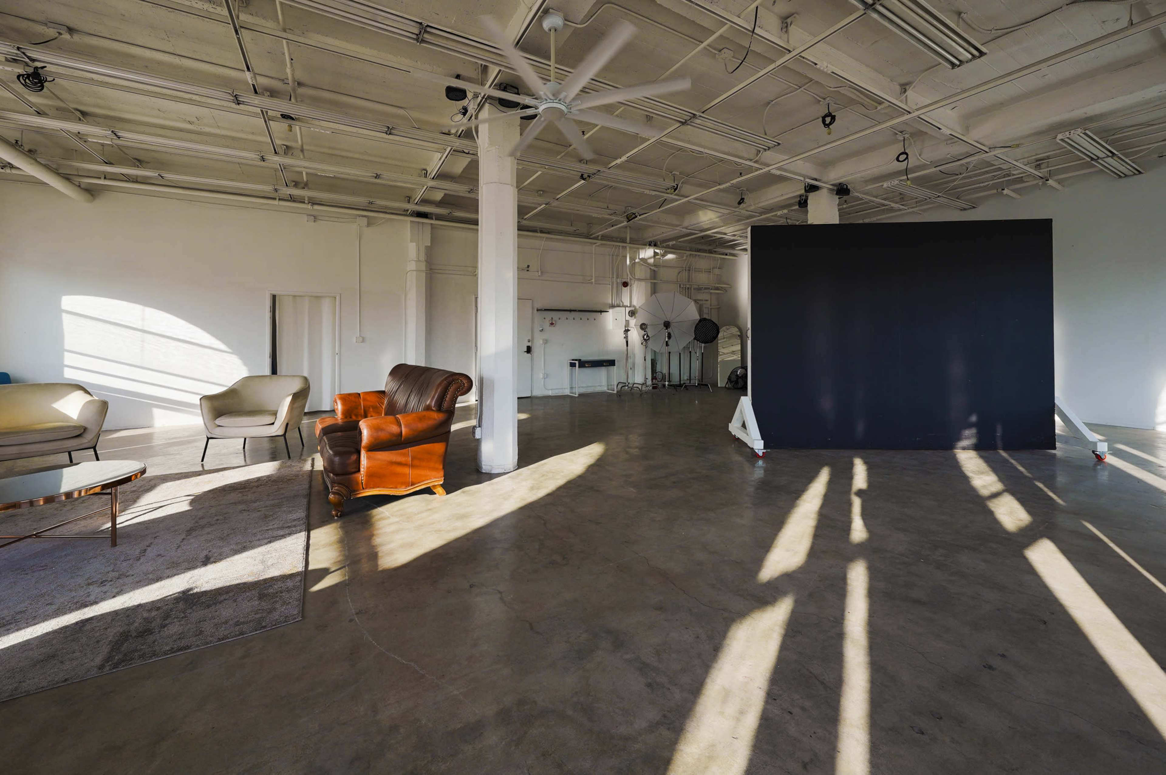 A spacious, well-lit studio features a large black backdrop, a brown leather armchair, a light-colored sofa, and concrete flooring with sunlit shadows.