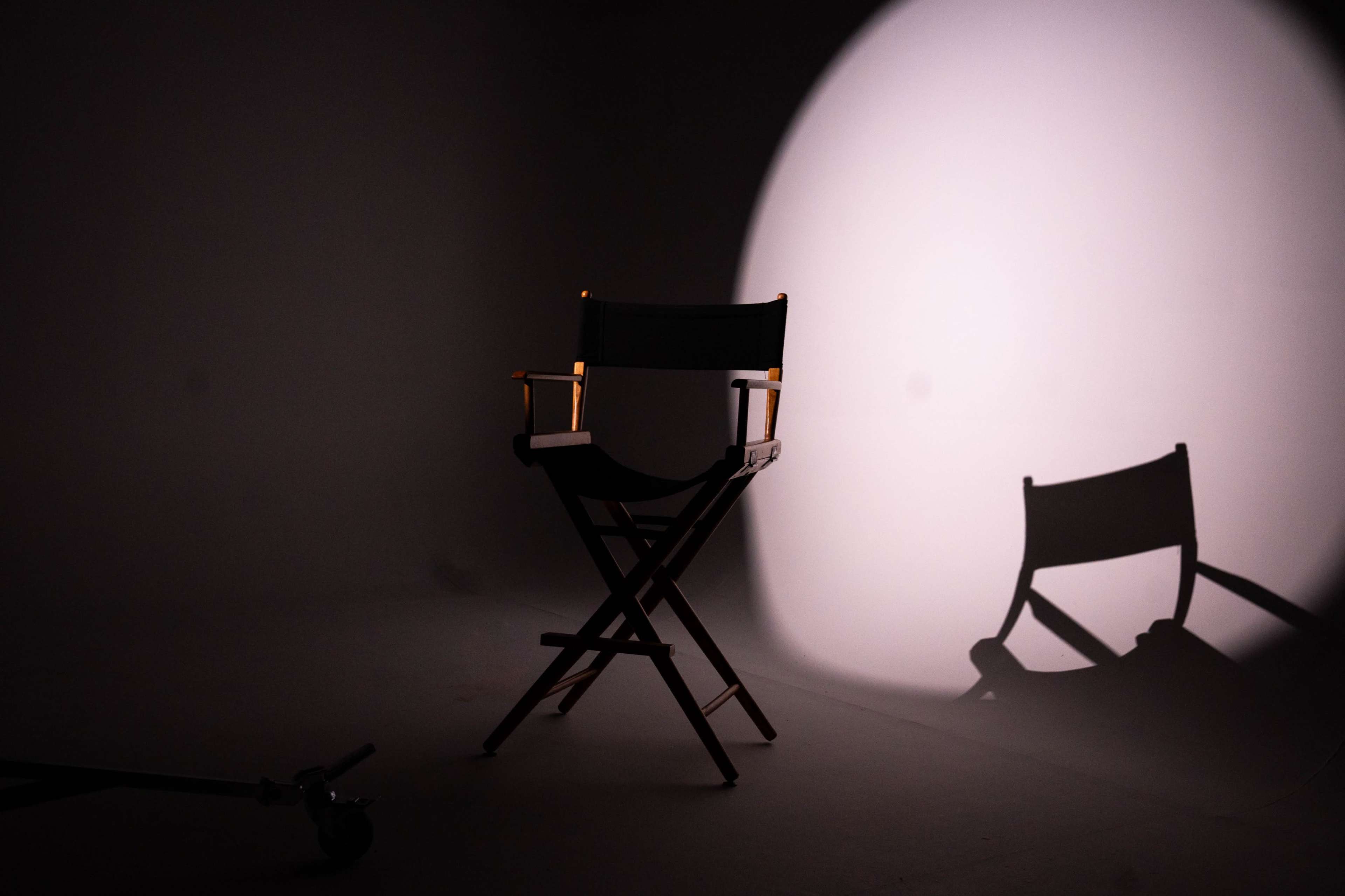 A director's chair stands alone in a dimly lit space, casting a shadow on the ground.